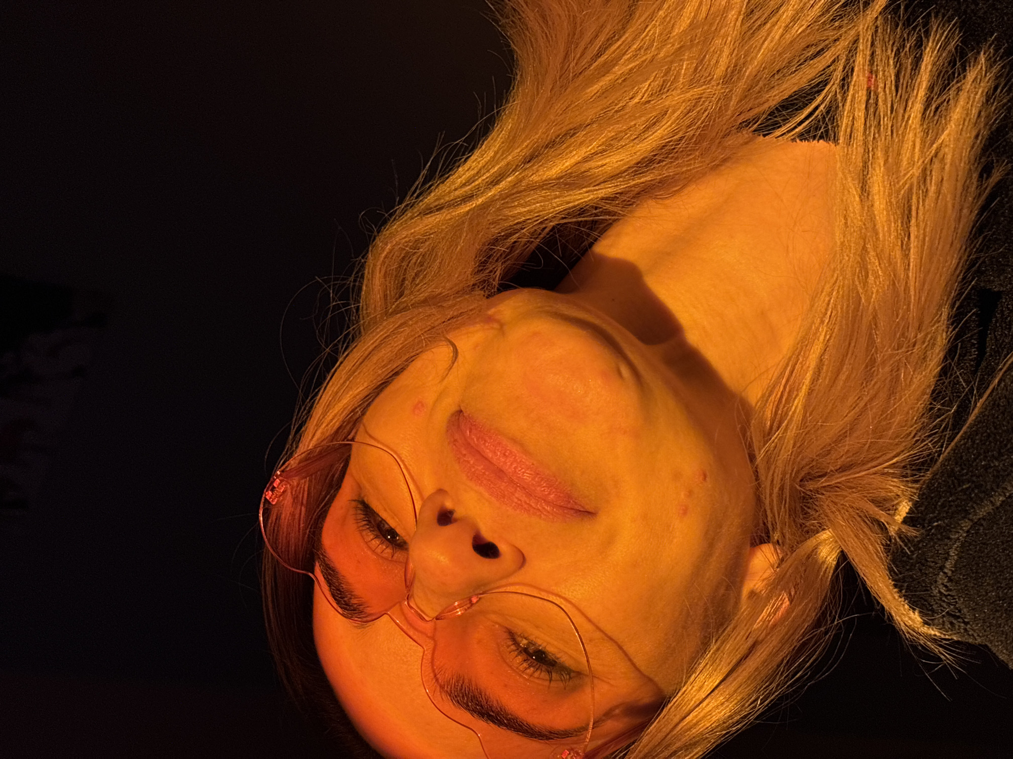 A close-up, vertically inverted image presents a woman with light-colored hair and wearing pink heart-shaped glasses. The lighting is predominantly orange, casting distinct shadows across her face and neck. The background is dark and indistinct. The location and city in Russia are not discernible from the image.