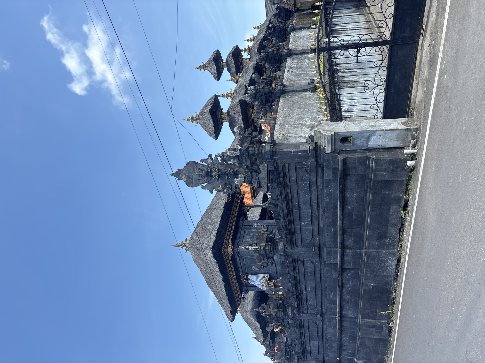 The image depicts a Balinese Hindu temple structure located in Kuta Selatan, Indonesia. The temple is constructed of dark stone, possibly volcanic rock, and features tiered pagoda-like roofs adorned with thatch. Decorative elements and carvings are visible on the stone structure. Adjacent to the temple, there is a wrought-iron gate. The temple sits alongside a paved road. Overhead, power lines intersect the clear blue sky with scattered clouds. The photograph appears to be taken at ground level, looking up at the temple.