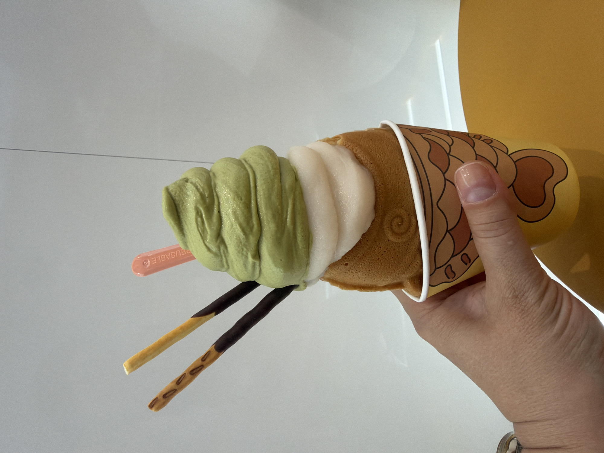 A hand is holding a taiyaki-shaped ice cream cone in a paper cup. The ice cream is layered with green matcha on top and white underneath. Two chocolate-dipped biscuit sticks are inserted into the ice cream, and a small pink plastic spoon is also present. The taiyaki itself appears to be a golden-brown pastry with a swirled pattern. The background is a plain, light-colored wall and a yellow surface. There is no indication that this image was taken in Prague, Czechia.