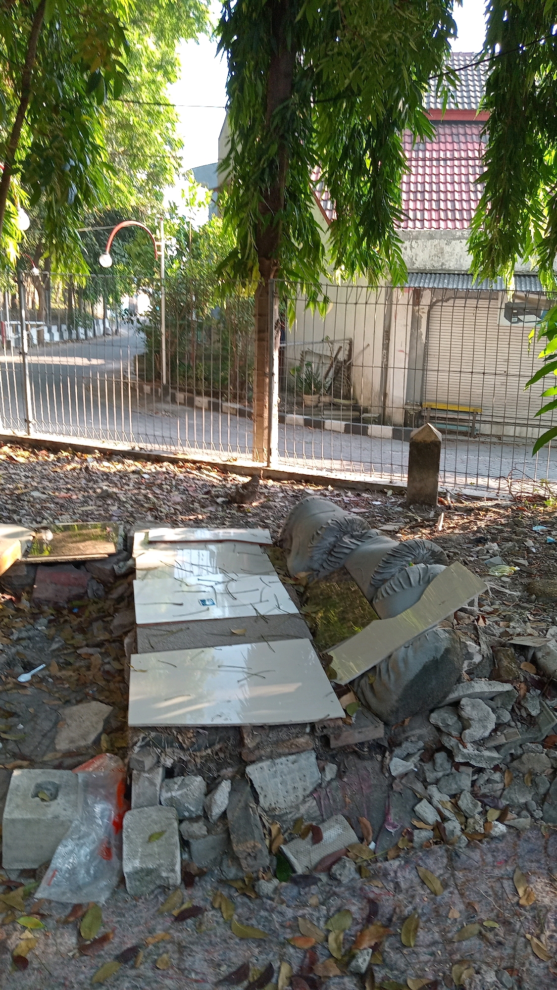 In Randuagung, Indonesia, a scene of disarray is depicted. In the foreground, broken concrete debris and fallen leaves litter the ground. Several white tiles are arranged on top of some of the rubble, with some appearing cracked. To the right, a large, grey, statue-like object lies on its side, its details obscured by shadows and debris. Behind this, a weathered fence separates the immediate area from a paved path and a street, suggesting a public or semi-public space. The background features a building with a red tiled roof and what appears to be a garage door, further indicating a residential or commercial area. Lush green trees with long, drooping leaves frame the scene, their branches reaching towards the bright sky. The overall impression is one of neglect and abandonment within a tropical environment.