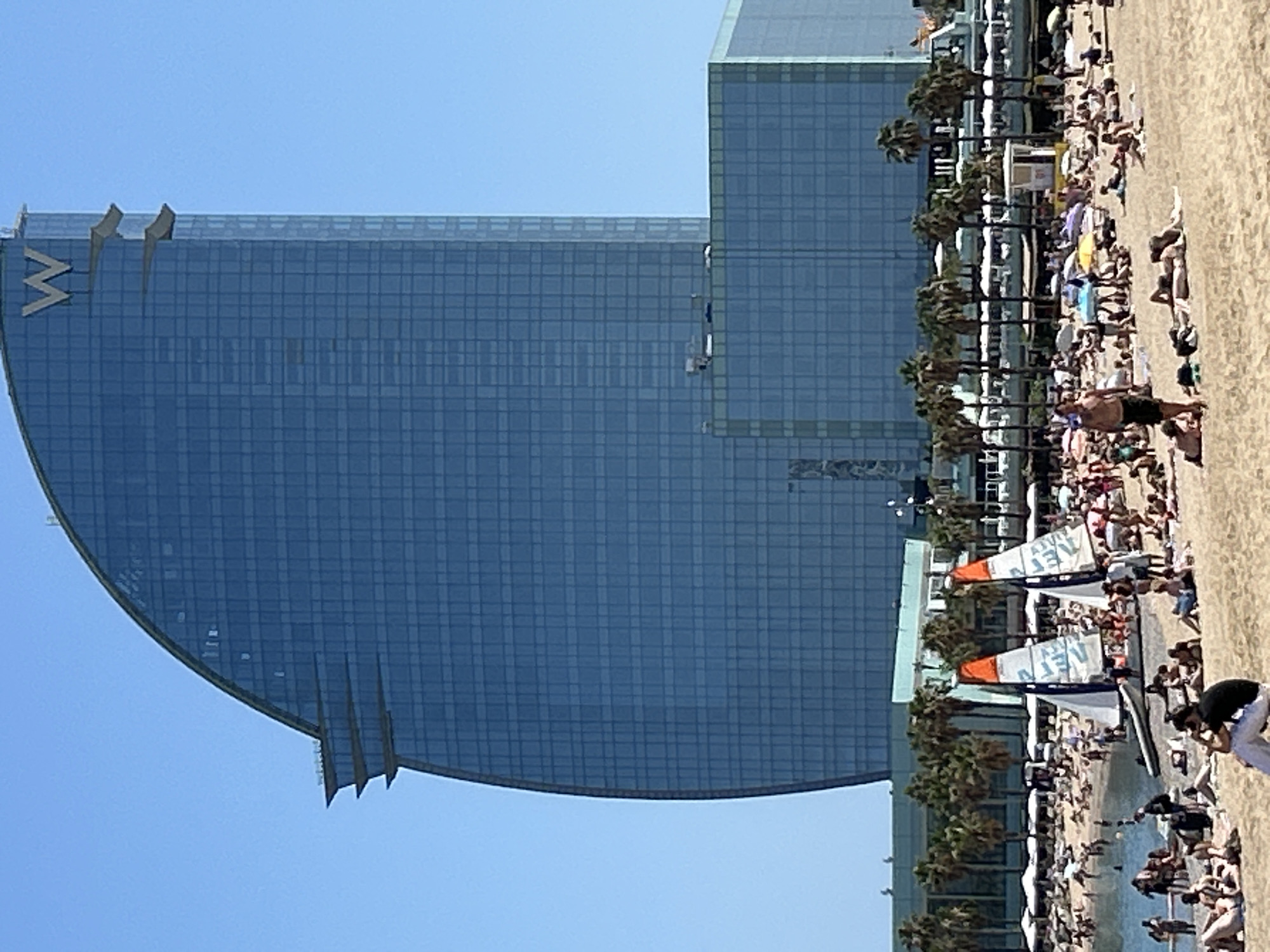 The image depicts a sunny day at Barceloneta Beach in Barcelona, Spain. A large, modern, curved glass building, identified by a prominent "W" logo at the top, dominates the background. The building is a hotel, likely the W Barcelona. In the midground, palm trees line the edge of the beach, behind a row of beach umbrellas and chairs. The beach itself is crowded with people sunbathing, walking, and interacting. Two small sailboats with orange and white sails are visible in the water. The sea is calm and blue, and the sky is clear. The overall atmosphere is one of a busy, popular tourist destination.