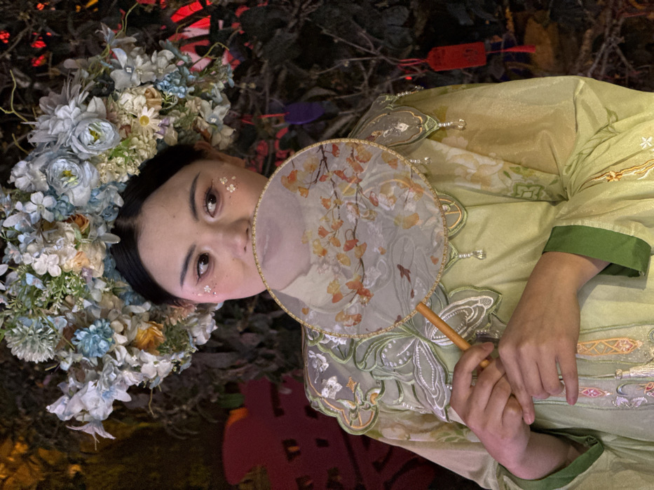 An East Asian female stands in a dimly lit setting within Yuzhong District, China. She wears a prominent floral headpiece composed of artificial flowers in shades of light blue, white, cream, and green foliage. Her visible facial features include eye makeup with winged eyeliner and iridescent glitter applied beneath the outer corners of her eyes. She is dressed in a light green traditional-style garment, likely a Hanfu, featuring intricate green and white embroidery, particularly around the collar which exhibits a scalloped design and hanging decorative beads. She holds a circular, translucent fan with a golden border and a wooden handle, positioning it to obscure her lower face. The fan's surface is adorned with an artistic depiction of autumnal-colored leaves on branches and two small fish. Her hands are clasped around the fan's handle. The background consists of dark, dense foliage, with blurred red, purple, and blue decorative elements, including a distinct red tag with yellow characters positioned on the right side.