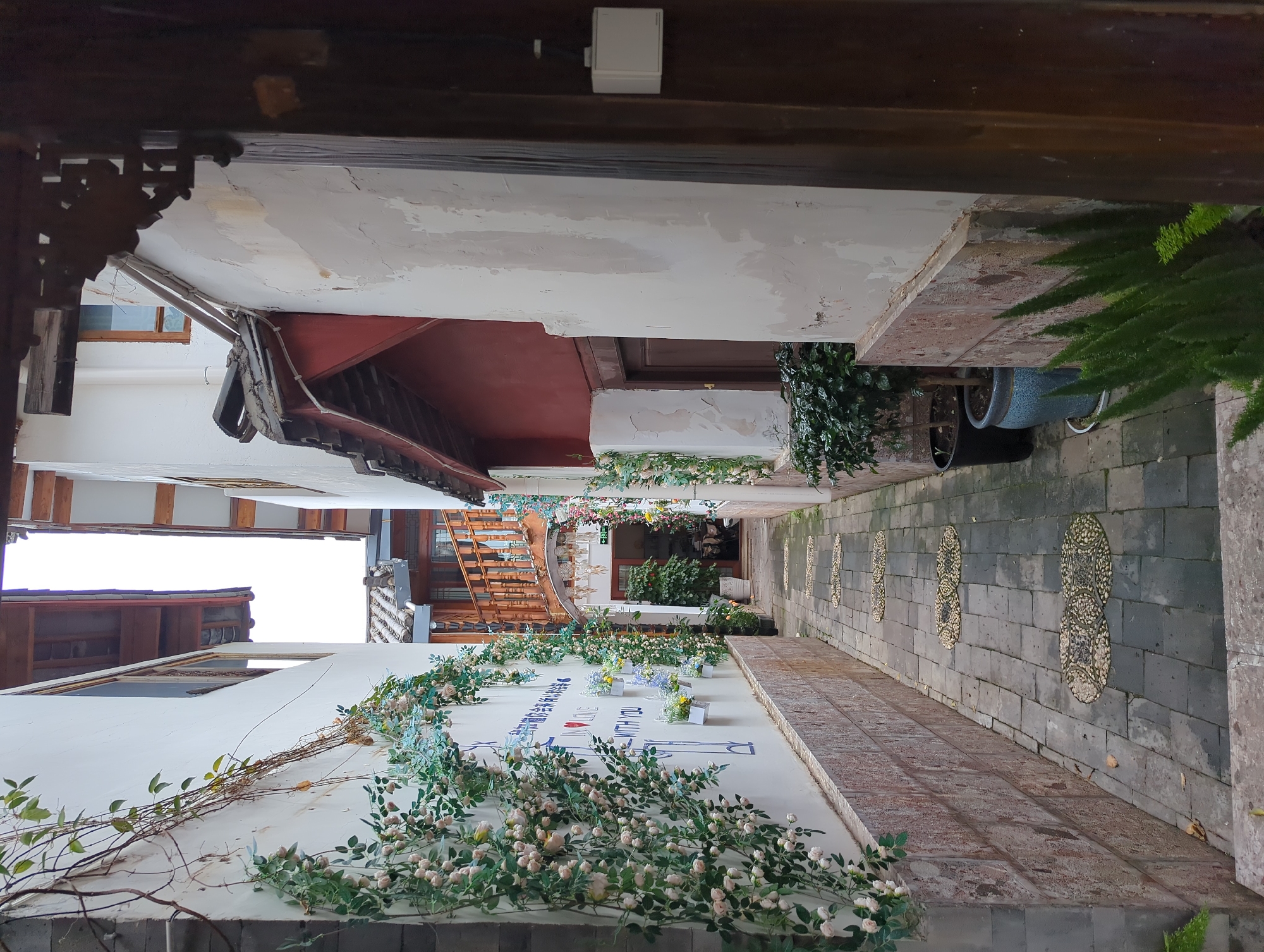 This image depicts a narrow alleyway in a traditional Chinese setting, likely in Gucheng District, China, given the architectural style. The alley is lined with buildings featuring white walls and dark wooden eaves. The ground is paved with stone tiles, and decorative elements like pebble mosaics are visible on the walls. Lush greenery, including climbing plants with delicate white flowers, adorns the walls, adding a touch of natural beauty to the scene.
In the foreground, a white wall is covered with these floral arrangements, with a vine gracefully trailing across it. There appears to be some text and decorative elements on the wall, possibly part of a mural or signage, featuring blue lettering and a heart symbol. In the background, more buildings and architectural details, including wooden staircases and doorways, can be seen, suggesting a complex of shops or residences. The scene is devoid of people, creating a sense of peacefulness and solitude.
The time of day appears to be overcast, as there are no strong shadows, and the light is diffused. The overall atmosphere is serene and picturesque, highlighting the charm of traditional Chinese architecture and its integration with nature. The decorative floral elements suggest an occasion or a deliberate aesthetic choice to enhance the visual appeal of the alleyway.