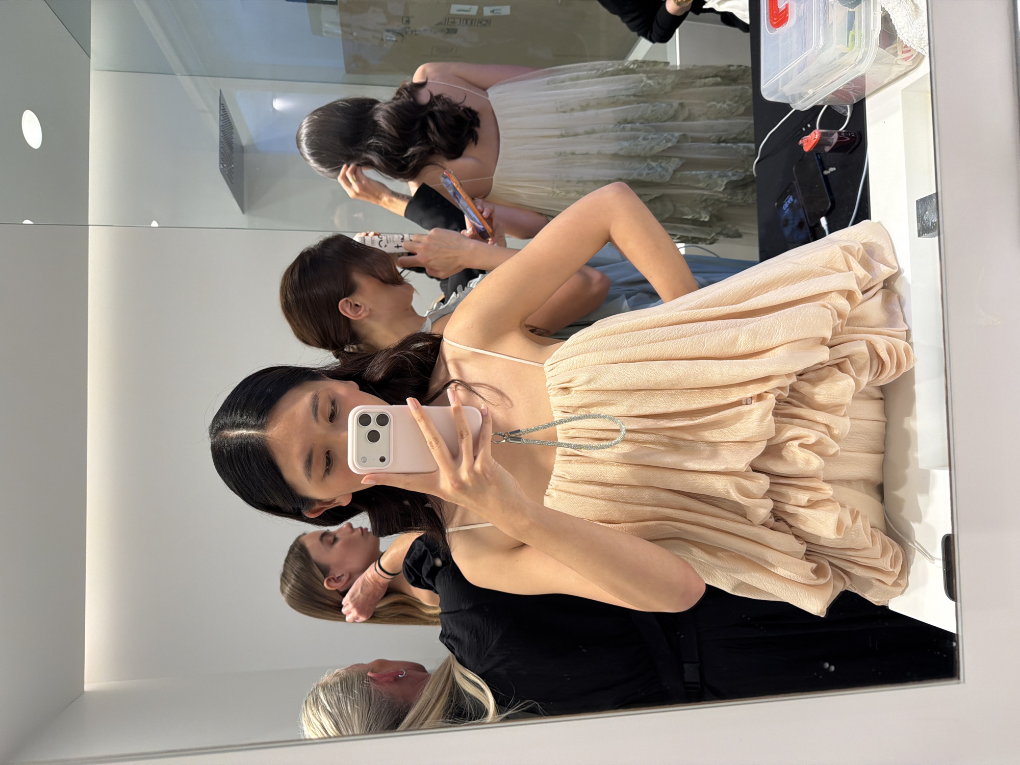 The image appears to be a selfie taken in a mirror. The person taking the photo is an Asian woman wearing a light peach-colored dress with a ruffled or draped design. She is holding a smartphone in front of her, using it to capture the image. Her black hair is straight and parted in the middle. In the reflection, there are three other women behind her. One woman with light brown hair and another with dark brown hair appears to be looking at their phones or cameras. The third woman has gray hair and is wearing a black top. The scene takes place in what seems to be a dressing room or backstage area, with visible makeup products and other personal items near the mirror's edge. Based on additional information provided, the location is Ramatuelle, France.