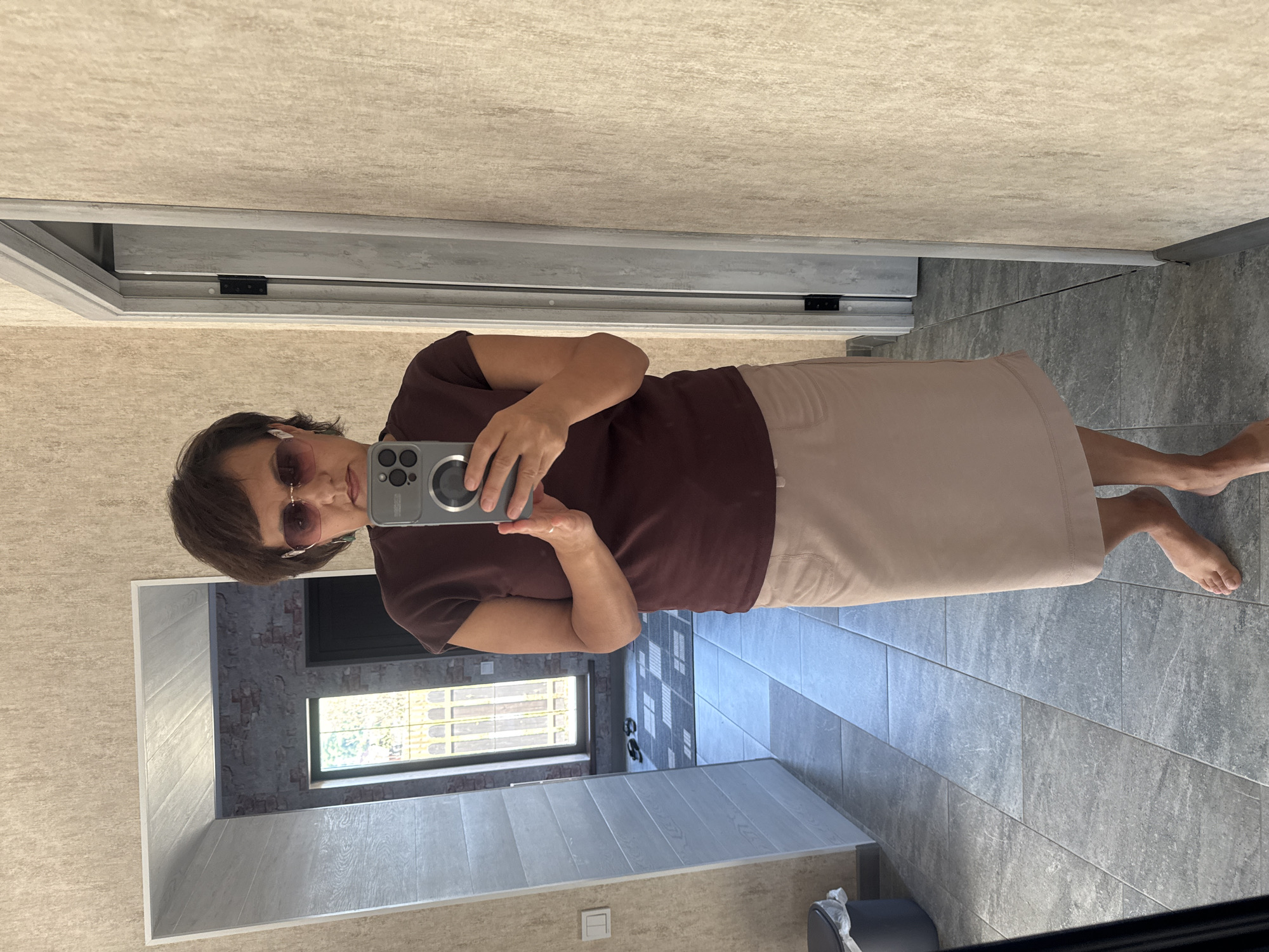 A woman with short brown hair and sunglasses is taking a mirror selfie in what appears to be a hallway or entryway of a building. She is wearing a dark maroon short-sleeved t-shirt and a light beige knee-length skirt. She is holding a smartphone in her right hand, pointing it towards the mirror. The background shows a door, textured walls, and a tiled floor. Through a doorway in the reflection, a window with a view of greenery and a brick wall is visible. The overall environment suggests an indoor residential or commercial space. The location is stated to be in the city of Soloneshnoye, Russia.
