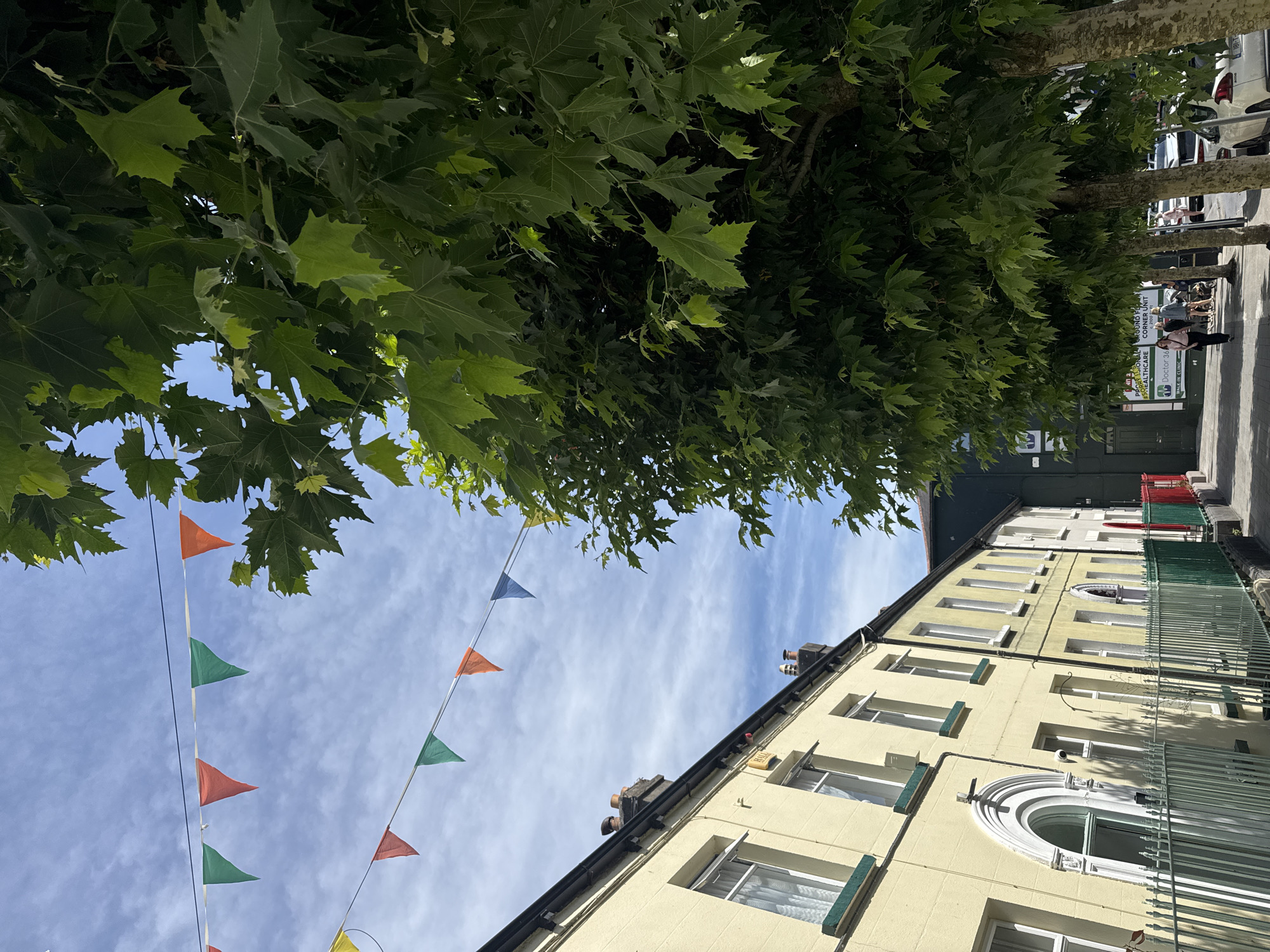 This image depicts a street scene in Clonmel, Ireland. A row of light yellow buildings with white-trimmed windows lines the left side of the street. Some buildings have ornate architectural details around the doorways. A green metal fence runs along the sidewalk in front of these buildings. Across the street, a large tree with lush green leaves dominates the right side of the frame. The tree's canopy partially obscures the sky, which is a bright blue with wispy white clouds. Strands of colorful bunting, consisting of orange, green, red, and blue triangular flags, are strung across the street, suggesting a festive occasion. The sidewalk continues along the right side, where several people are walking. Some parked cars are visible further down the street. A green building with signage is also present on the right.