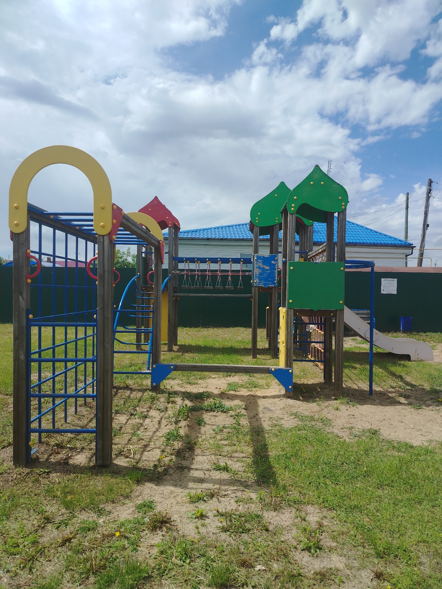 The image shows a children's playground in Klyuchi, Russia, on a cloudy day. The playground features a climbing structure with red rings, a blue ladder, and a yellow arch. To the right, there is a green roofed structure with a slide. In the background, a teal fence encloses the area, and a blue-roofed building is visible beyond. The ground is a mix of sand and grass. No people are present in the image.