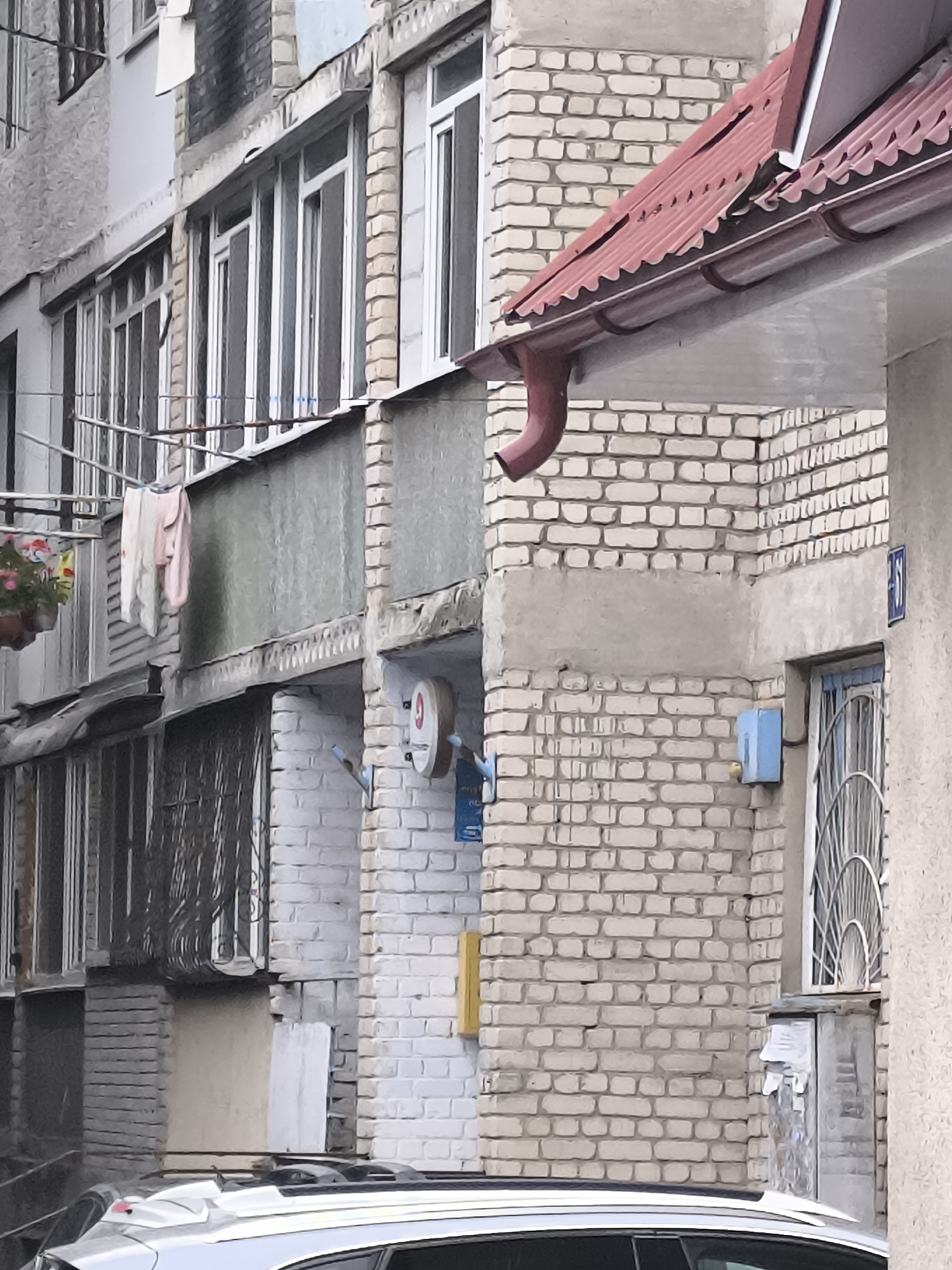 The image depicts the exterior of a brick building, likely an apartment complex, in Bălți, Moldova. The building features multiple floors with windows, some of which are covered with metal grates. A red, corrugated metal roof with a gutter system is visible at the top right, with a downspout extending downwards. Laundry, including a pink and white garment, hangs from a clothesline on a balcony on the left side of the building. A potted plant with pink flowers is also visible on this balcony. A blue electrical box is mounted on the brick wall to the right of center. A metal grate covers a window above the blue box. A blue sign with the number "61" is affixed to the wall above the window. The bottom of the image shows the rear of a silver car. The overall impression is one of a typical urban residential building in a state of moderate disrepair, with elements of daily life like laundry drying.