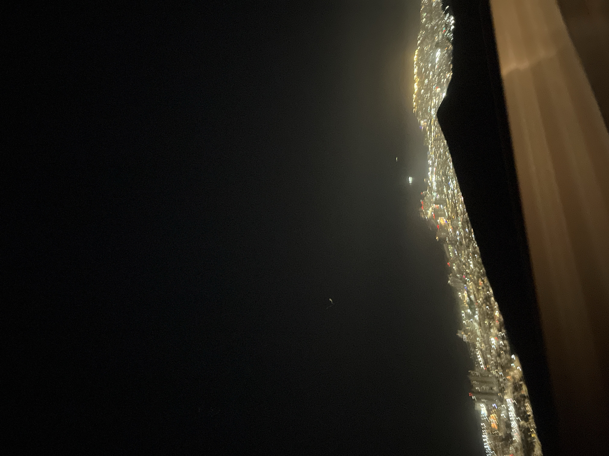 The image shows a dark night scene, likely taken from a high vantage point. On the right side, a blurry, vertical strip of bright, white and yellow lights indicates a city skyline. To the left of the skyline, the scene is predominantly dark, suggesting a body of water or open space. The edge of a window frame is visible on the extreme right, with a hint of a curtain. Overall, the image depicts an urban cityscape at night, viewed from an elevated position.
