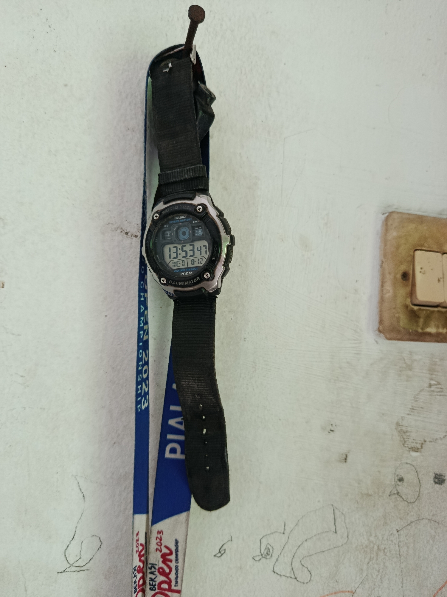 The image shows a digital Casio watch hanging on a nail against a textured white wall. The watch displays the time as 13:53:47 and the date as Wednesday, August 12. The watchband is black fabric. Below the watch, two blue lanyards with text printed on them are also hanging on the nail. The text on the lanyards appears to be related to a "CHAMPIONSHIP 2023" and "PIALA OPEN" in what looks like Bahasa Indonesia. The wall has some faint scratches and a light switch with two toggle buttons is visible to the right of the watch. There are also some faint pencil drawings on the wall, including what appears to be a bird and abstract shapes. The environment suggests an indoor setting, possibly a home or a casual establishment in Wanasari, Indonesia, given the linguistic cues.