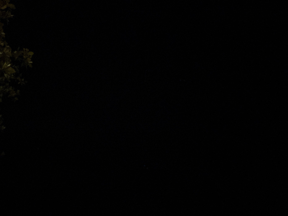 The image is predominantly black, with a minimal amount of dark, out-of-focus green foliage or leaves dimly discernible in the upper right quadrant. No individuals, identifiable subjects, distinct objects, specific actions, or interactions are visible. The environment appears extremely dark, consistent with low light conditions. The precise location within Ban Ko Li Pe, Thailand, cannot be determined from the visual information provided.