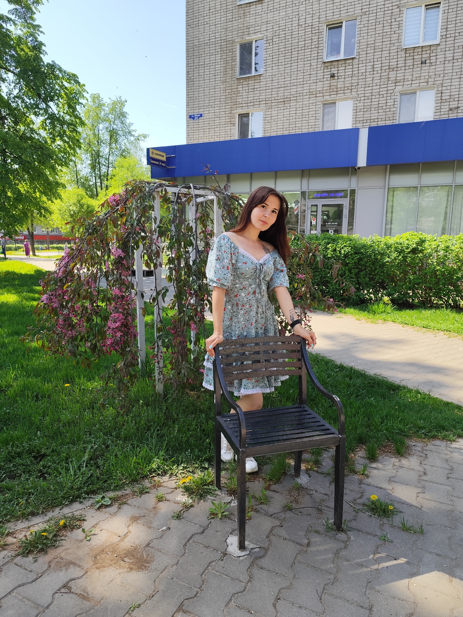 A medium shot captures a woman in a floral dress standing behind a black metal chair on a brick pathway with patches of grass. She has long brown hair and is leaning slightly on the chair with her left hand. A flowering tree with pink blossoms is to her left. Behind her, there is a building with a blue awning and signs in Russian, with an address marker reading “ул. Мира 20”. A hedge separates the pathway from the building. The location is Chaikovsky, Russia.