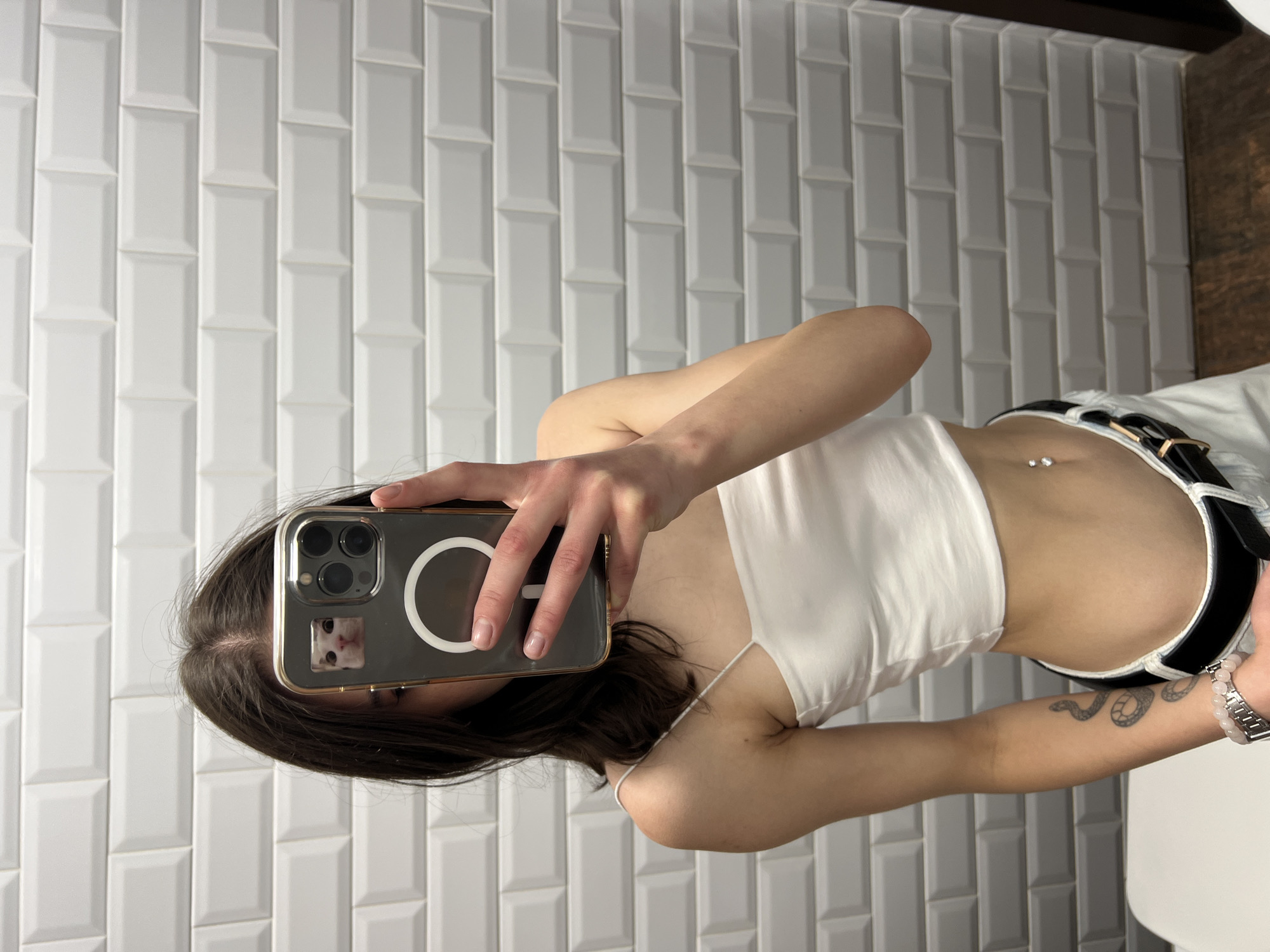 A young woman is taking a mirror selfie in a room with white subway tile walls. She is wearing a white crop top, white pants, and a black belt. She has a belly button piercing and a snake tattoo on her left arm. She is holding up her phone, which has a sticker of a cat on the back. The image is taken from the chest down. The floor is a dark wood.