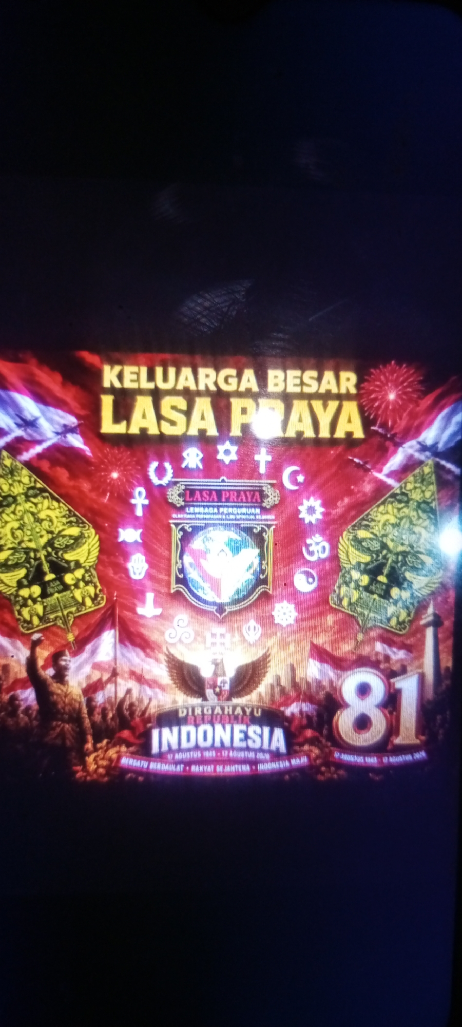 The image is a screenshot of a large banner or screen display, likely from an event in Cemani, Indonesia. The display features a patriotic theme, celebrating "Dirgahayu Republik Indonesia" (Happy Birthday Republic of Indonesia). Dominating the center is the number "81" in large, golden numerals, suggesting the 81st anniversary.
The banner prominently features the text "KELUARGA BESAR LASA PRAYA" in large, bold, yellow lettering. Below this, the name "LASA PRAYA" is repeated within an emblem, accompanied by the words "LEMBAGA PERGURUAN." The emblem itself contains abstract imagery and symbols.
Scattered around the emblem are various symbols that appear to represent different religions or spiritual beliefs, including what looks like a crescent moon and star (Islam), a cross (Christianity), a Star of David (Judaism), and symbols resembling Buddhist and Hindu iconography.
The lower half of the display depicts a stylized scene of Indonesian independence. A figure in military attire, with a raised fist, stands at the forefront, symbolizing a leader or revolutionary. Behind him, a crowd of people waves Indonesian flags, which are red and white. The Garuda Pancasila, the national emblem of Indonesia, is also visible.
The overall aesthetic is vibrant and celebratory, with red, white, and gold as dominant colors. The display seems to be positioned outdoors or in a large venue, possibly decorated with fireworks and other festive elements. The lighting suggests it might be nighttime or an indoor event with dramatic lighting. The context of Cemani, Indonesia, places this display within a specific geographical and cultural setting.