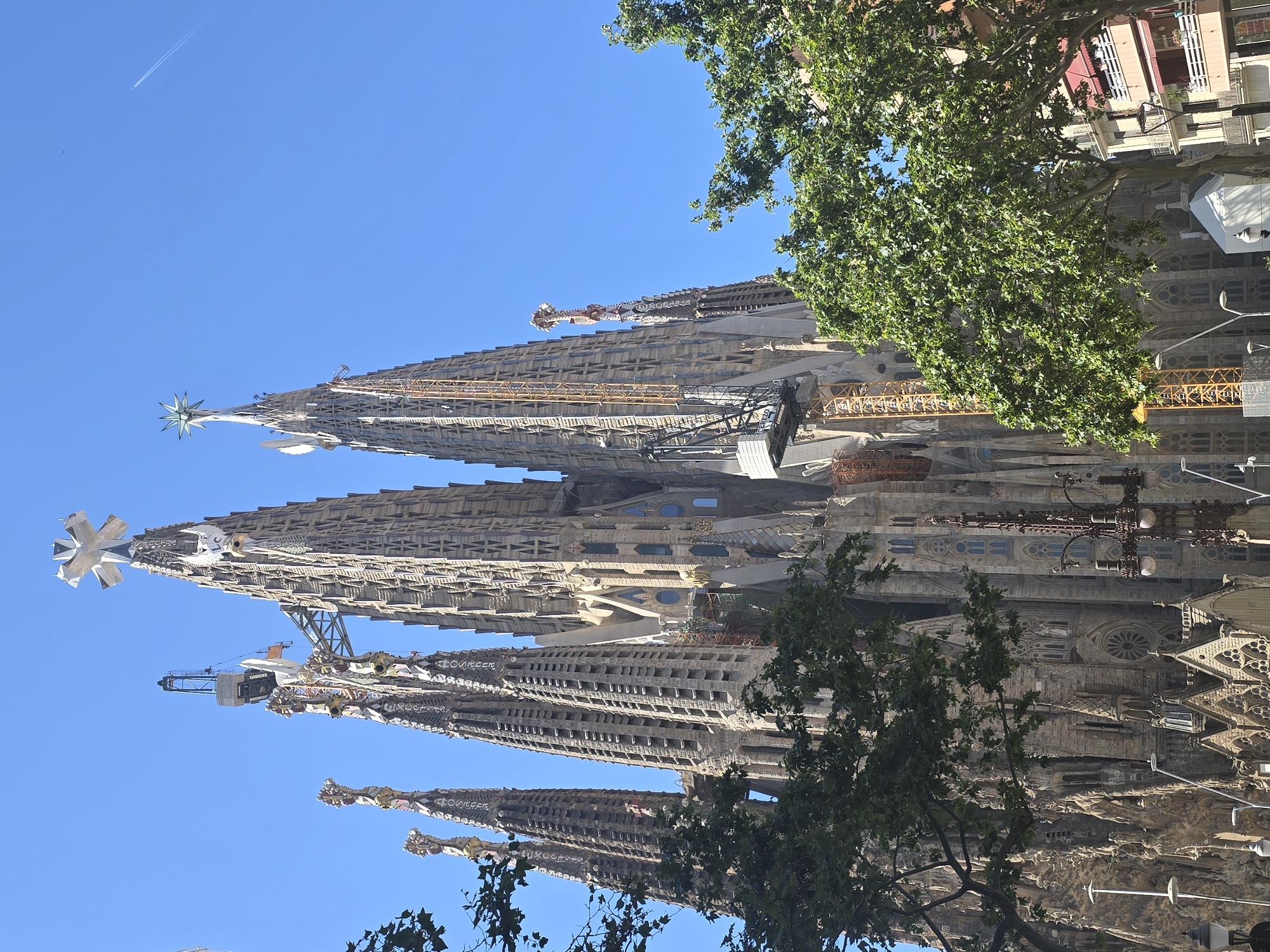 The image captures a low-angle, full-frame shot of the Sagrada Familia basilica in Barcelona, Spain, under a clear blue sky. The iconic, unfinished Gothic-style basilica dominates the frame, with its intricate stone facade and towering spires reaching towards the sky. Several cranes, including a large yellow construction crane and a smaller grey one, are visible against the structure, indicating ongoing construction. Lush green trees partially obscure the lower left and right portions of the basilica, framing the grand architectural marvel. In the background, a few buildings with balconies and red awnings are visible, along with a distant contrail in the sky, suggesting air traffic. The overall scene conveys a sense of scale, complexity, and ongoing work on a world-renowned landmark.