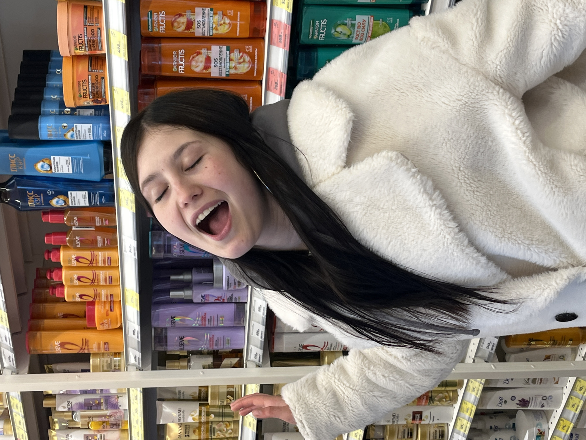 A young woman in a white, fluffy jacket stands in a store aisle in the city of Chaikovsky, Russia. Shelves are stocked with hair care products like Garnier Fructis, Elseve, Pantene, and Dove. The woman is photographed from an unusual angle, with her head tilted back and mouth open in a wide, expressive gesture. She has long, dark hair and is wearing hoop earrings. The focus is on her and the hair product selection.