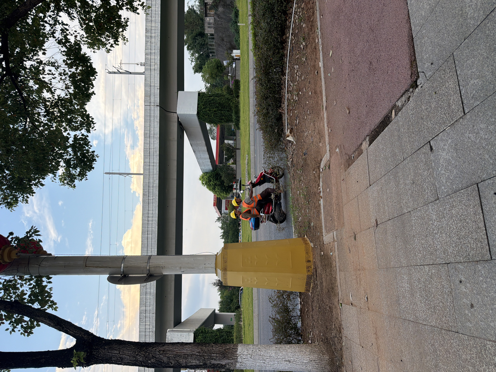 Two individuals wearing yellow hard hats and orange safety vests are riding on a red scooter. They are positioned on a paved road, with a sidewalk in the foreground. Behind them, a large yellow structure with intricate patterns stands next to a streetlight pole. Overhead, a concrete overpass, likely part of a highway or railway, spans across the scene, supported by large pillars. Trees and green vegetation are visible in the background. The sky is partly cloudy. The location is identified as Jiangjin, China.