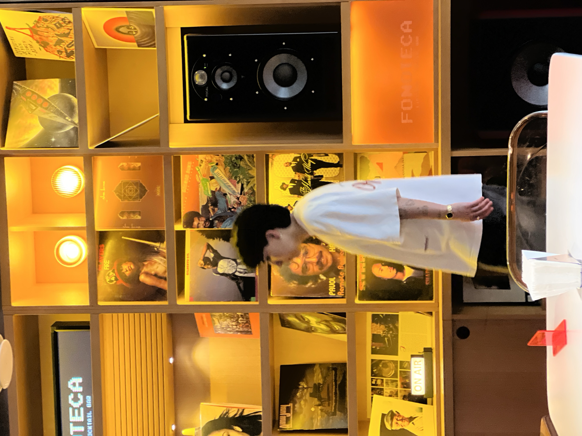The image shows a man standing in front of a shelf full of records. The shelf is made of wood and has a light behind it, illuminating the records. The man is wearing a white shirt and has dark hair. The records on the shelf are a mix of genres, including classical, jazz, and rock. A Focal brand speaker is visible on the shelf. The location is possibly a bar or restaurant in Yerevan, Armenia, named "FOMECA."