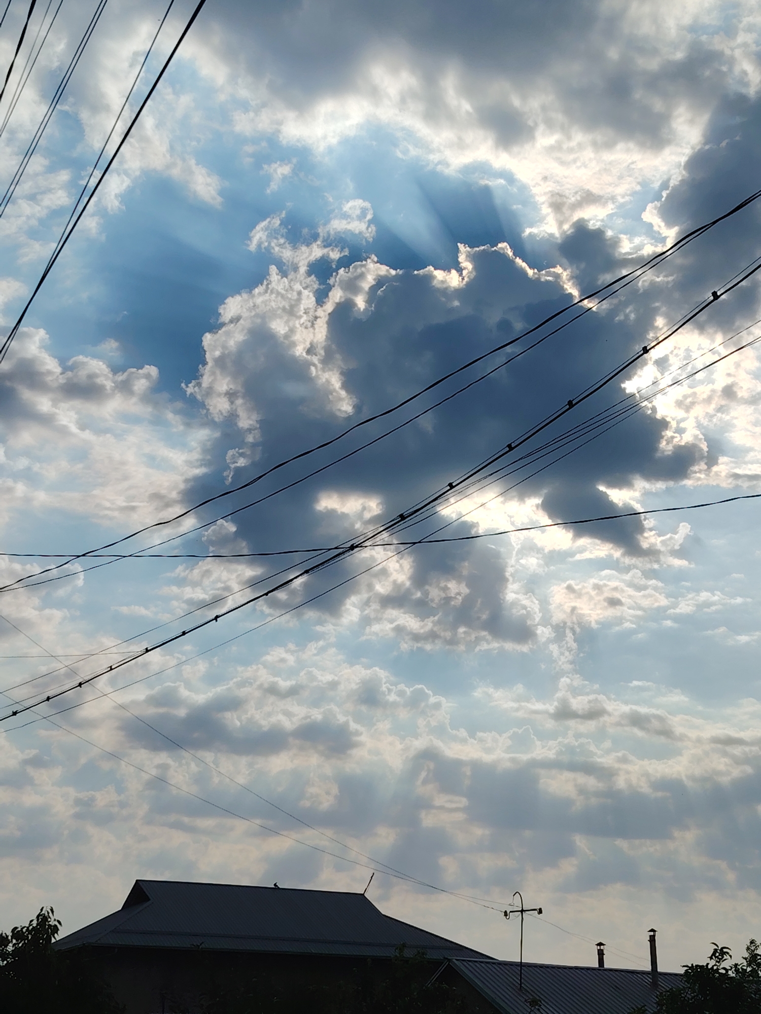 The image depicts a view of the sky with clouds and sunbeams, partially obstructed by overhead power lines and silhouetted rooftops of buildings. The sky is partly cloudy, with dramatic sunbeams (crepuscular rays) piercing through gaps in the clouds, indicating a late afternoon or early morning sun. Several dark power lines crisscross the frame horizontally. The lower portion of the image shows the dark silhouettes of building rooftops, including what appears to be a house with a pointed roof and chimneys. The setting is a residential area in Tavaksay, Uzbekistan. No people are visible in the frame.