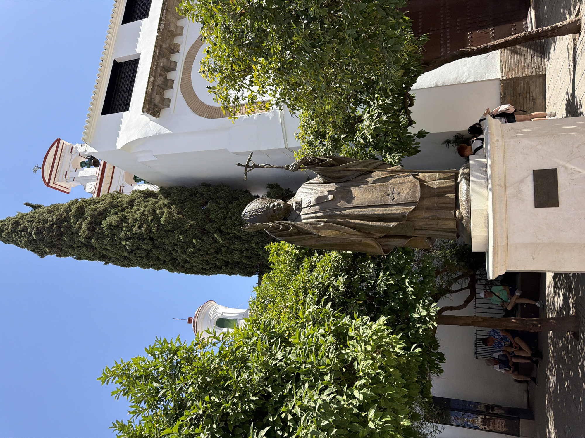 The image depicts a sunny day in what appears to be a plaza or courtyard in Sevilla, Spain. The central subject is a bronze statue of a robed figure, possibly a saint or historical personage, holding a cross. The statue is positioned in front of a white building with architectural details suggesting a church or historical structure, including arched windows and decorative molding. Lush green trees and foliage frame the scene, with sunlight dappling through the leaves, indicating a warm, clear day.
In the background, several people are visible. Some are standing near the building, while others are seated on benches beneath the shade of a tree. One person is leaning against a wall, and another is seated on a low wall, looking down. The presence of people suggests a public space, possibly a tourist attraction or a gathering place.
To the left of the statue, a section of a bell tower is visible, with a bell hanging in an open alcove. The overall impression is one of a peaceful, sunlit day in a historical setting, with the statue serving as a focal point amidst the architecture and natural elements. No visible text is present in the image.