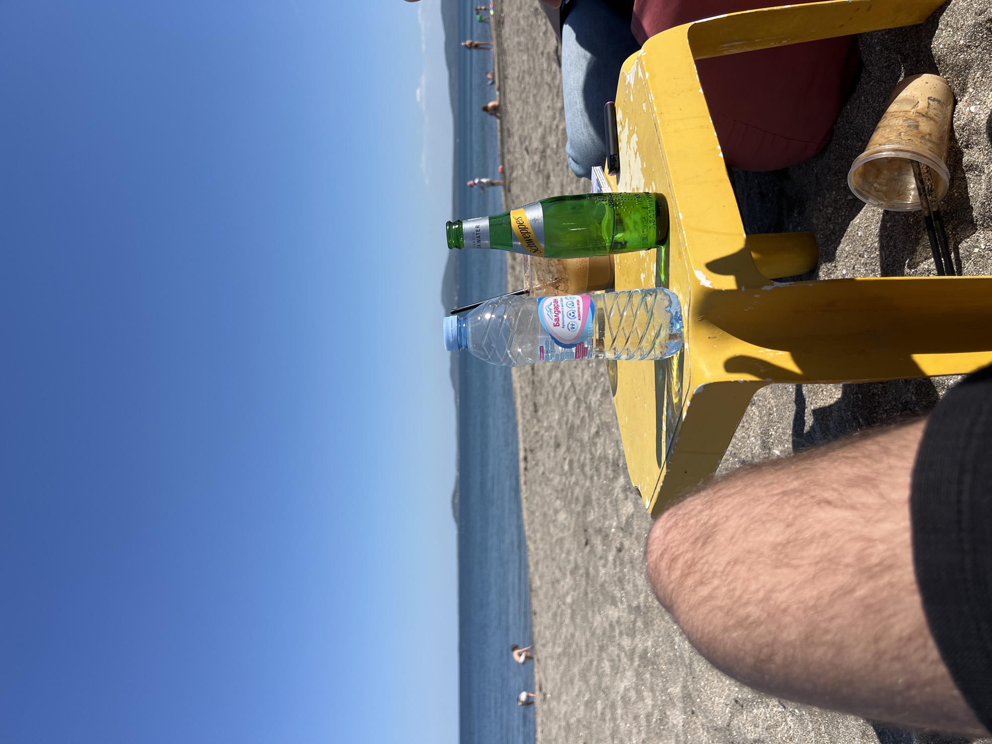 The image depicts a beach scene in Burgas, Bulgaria, featuring a yellow plastic chair on a sandy shore, with the Black Sea and blue sky in the background. A partially consumed bottle of Bulgarian mineral water and a Schweppes bottle are on the chair's surface. A disposable cup with chopsticks inside sits adjacent to the bottles. A person's leg is visible in the lower right corner of the frame, and several people are walking along the beach in the background.