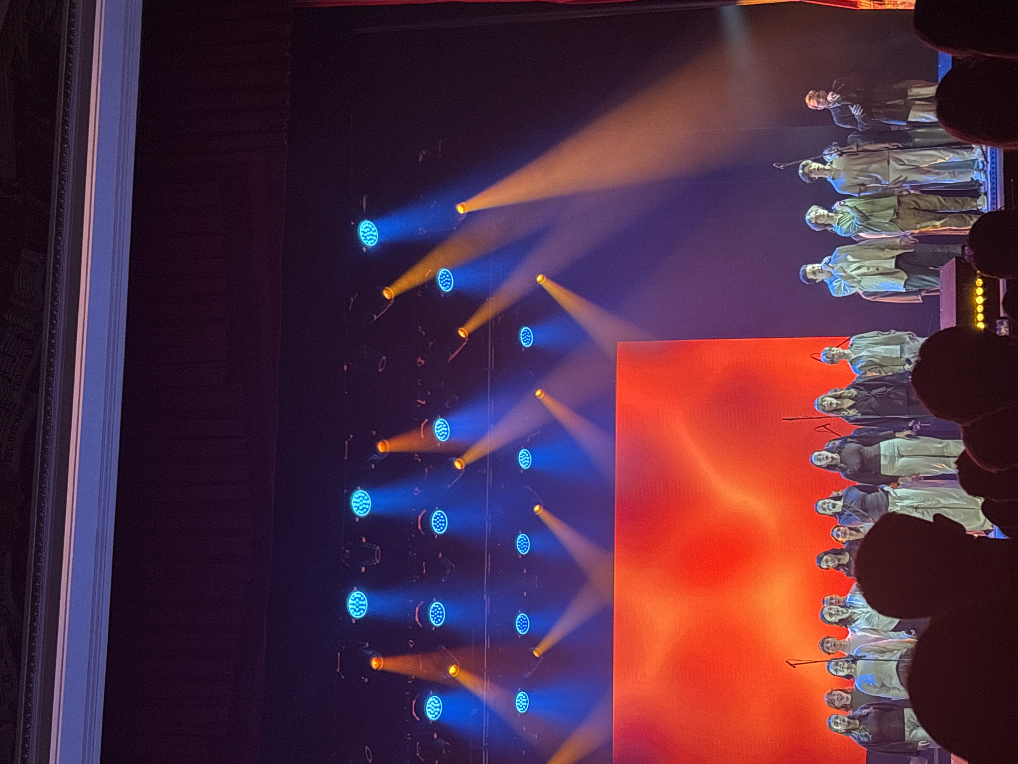 The image depicts a performance on a stage in Perm, Russia. A row of performers in various outfits stands in the background, illuminated by blue and orange lights. A large orange screen is partially visible on the right. Spectators are in the foreground. A decorated theater wall is on the left.