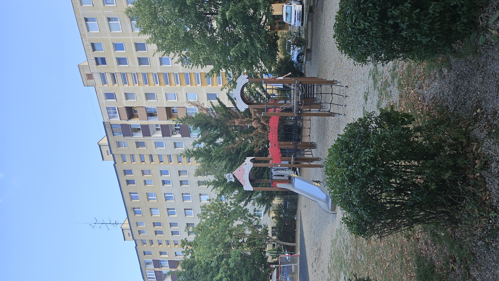 The image depicts a children's playground in front of a multi-story apartment building. The building, appearing to be of Soviet-era architecture, is light beige with dark brown accents around some of the balconies and window frames. A prominent antenna array is visible on the roof of the building. The playground features a slide, climbing nets, and a bridge-like structure. Trees are scattered around the playground and in front of the building. Two vehicles are parked near the building entrance: a silver car and a white van. The ground of the playground is covered with gravel and scattered leaves. The scene suggests an outdoor residential area. The location is identified as Bratislava, Slovakia.