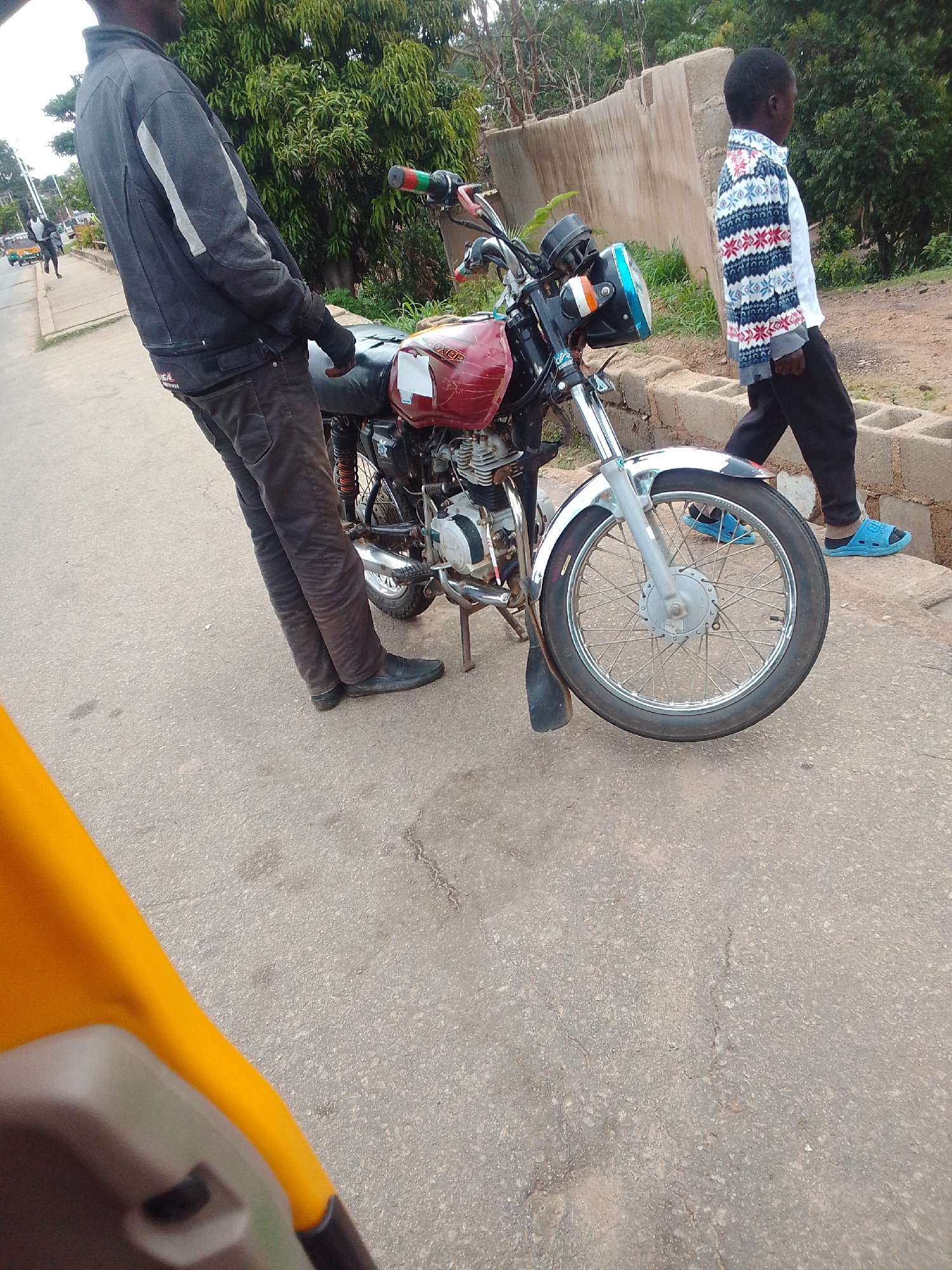 This photograph depicts a daytime street scene in the city of Jos, Nigeria. In the central mid-ground, an adult male with dark skin is standing, leaning against a red Bajaj Boxer motorcycle on an asphalt road. He is wearing a dark grey long-sleeved jacket with light grey vertical stripes on the outer arms, dark trousers, and dark slip-on shoes. His left hand rests on the motorcycle's black seat. The motorcycle features a red fuel tank with "BOXER" branding and some white tape, a chrome front fender, a prominent blue-tinted headlight with orange turn signal indicators, and its kickstand is engaged.
To the right and slightly behind the motorcycle, a young male, also with dark skin, is walking from right to left on a dirt or uneven concrete path. He is dressed in a long-sleeved white shirt with a patterned overlay of red, blue, and white geometric designs on the sleeves and shoulders, dark trousers, and bright blue slide sandals.
The background consists of stacked concrete blocks forming a low wall, a larger, light-colored concrete wall further back, and dense green vegetation, including trees and bushes. To the far left, a glimpse of a street extends into the distance, showing other vehicles, including what appears to be a tricycle (keke napep), and pedestrians. The immediate foreground, at the bottom left, is blurred and shows a yellow curved surface with a dark grey component, consistent with a close-up view from inside or immediately adjacent to another vehicle. The asphalt road surface exhibits minor cracks and wear.