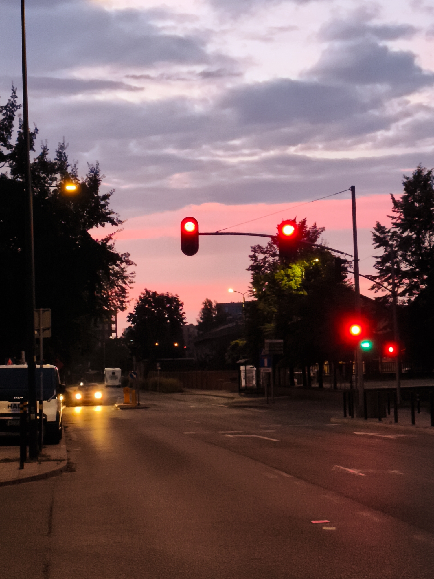 A twilight street scene captures a road in Gdańsk, Poland, as indicated by a sign on a white vehicle. The sky is a dramatic blend of pink and purple hues with scattered clouds, suggesting the end of a day. A traffic light in the foreground is illuminated with a red light, signaling a stop. Trees line the sides of the road, their silhouettes dark against the colorful sky. A white vehicle, possibly a van or minibus, is parked on the left side of the road, its headlights on and casting a yellow glow on the wet asphalt. The road itself appears to be asphalt, with painted lane markings faintly visible. In the distance, other vehicles and buildings are discernible, adding to the urban context. The overall impression is one of a quiet evening in a city, with the glowing traffic light serving as a focal point.