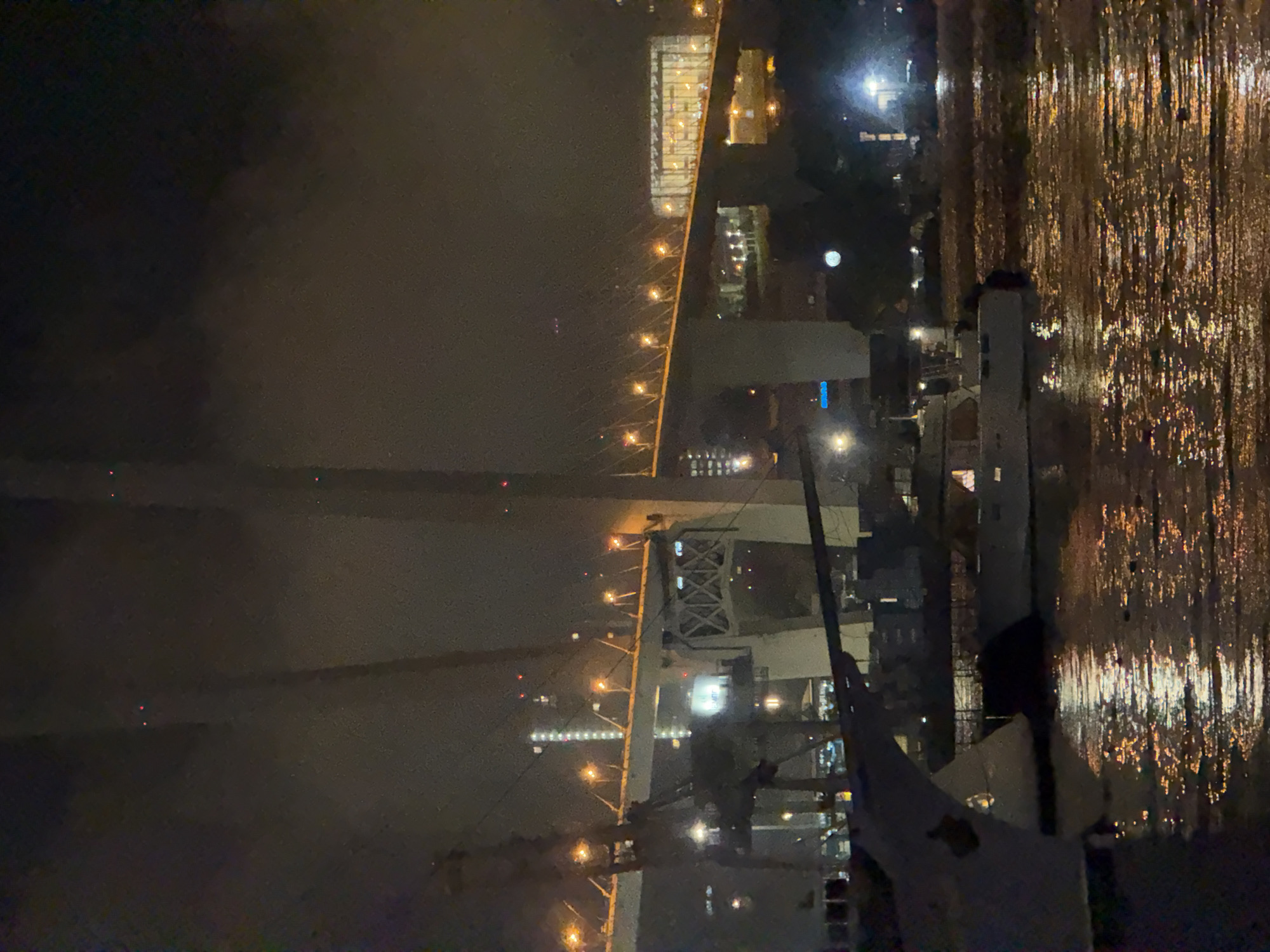 This is a nighttime photograph of a harbor in Vladivostok, Russia. In the foreground, several boats are docked on a body of water that reflects the lights of the city and the bridge above. The water appears choppy, with numerous highlights from the artificial illumination. Dominating the background is a large, illuminated bridge spanning the water, with numerous lights lining its structure. The bridge is supported by massive concrete pillars. Further in the background, city buildings are visible, some with lit windows, and the sky is dark and cloudy. The overall scene is one of urban nighttime activity with a focus on maritime infrastructure.