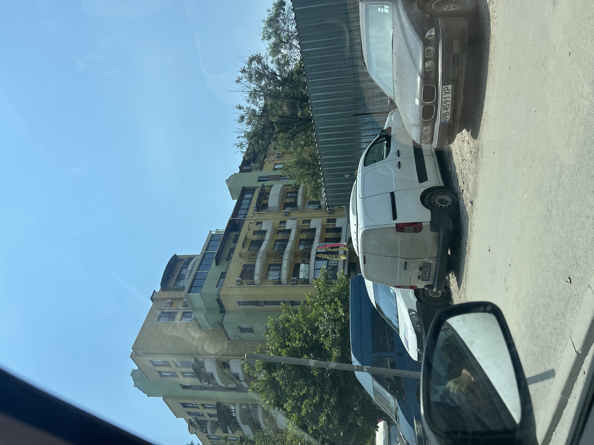This image captures a street scene in Plovdiv, Bulgaria. The main subjects are several parked vehicles, including a dark sedan with license plate "PB 4591 XP" and a white van. The setting is a city street, with a tall, multi-story building as the backdrop. The building is a mix of yellow and green, featuring numerous balconies and windows. Trees are visible in front of the building, suggesting a residential or mixed-use area. A corrugated metal fence runs along the edge of the road, separating the parked cars from the sidewalk or property behind.
The image appears to be taken during the daytime, under clear blue skies, indicating fair weather. The sun is casting shadows, particularly from the cars. One of the vehicles, the white van, has its rear door slightly ajar. Visible text includes the license plate on the sedan and potentially a sign on the building, though it is not clearly legible. The overall impression is a snapshot of everyday urban life in Plovdiv on a bright day.