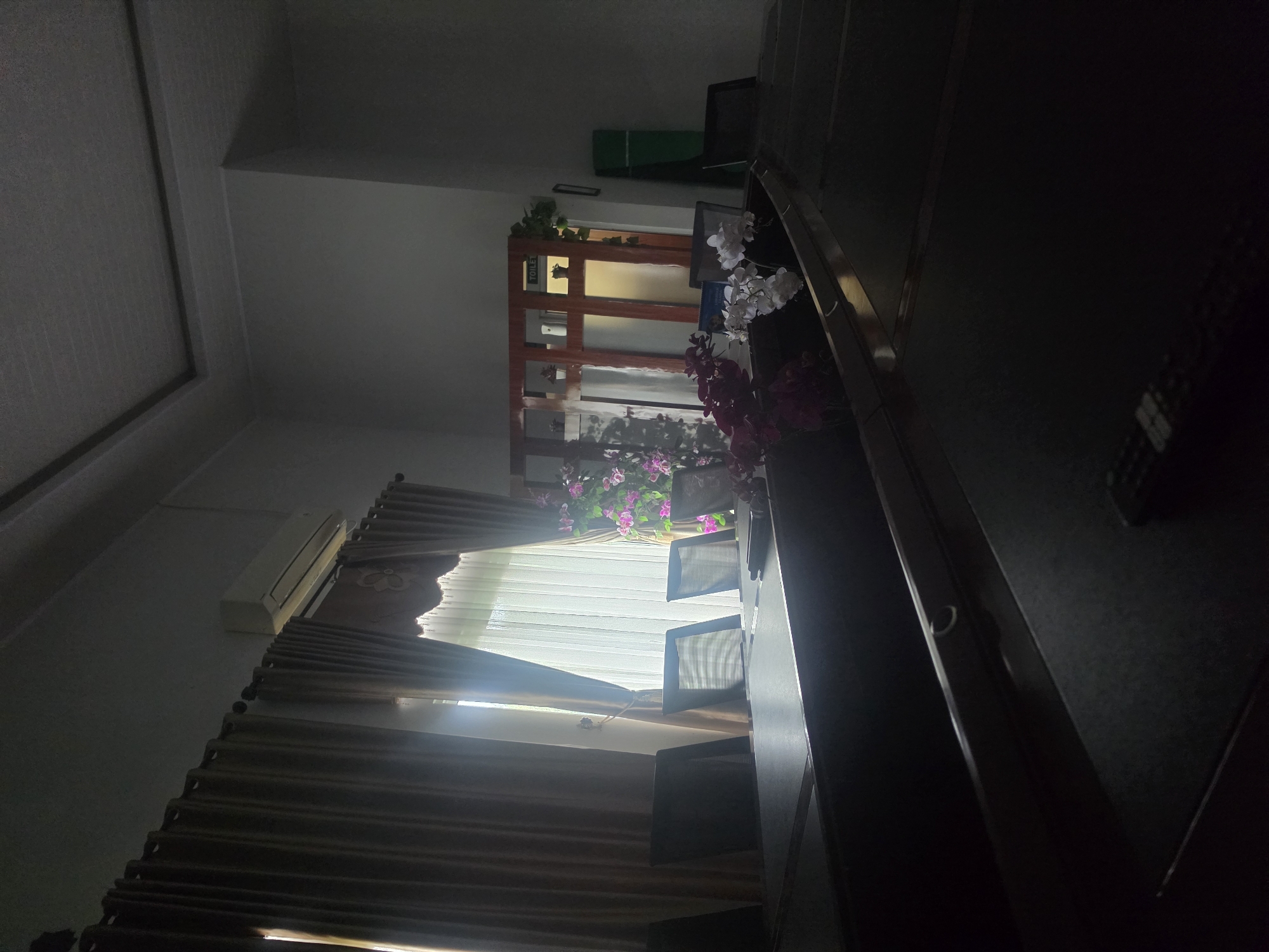 The image shows the interior of a room in Pintomakmur, Indonesia. A window with closed blinds allows light to enter. An air conditioning unit is mounted on the wall above the window. A wooden shelving unit is visible in the background, decorated with greenery and flowers. A staircase with a dark railing is positioned on the right side of the image. Several objects are scattered on the staircase.