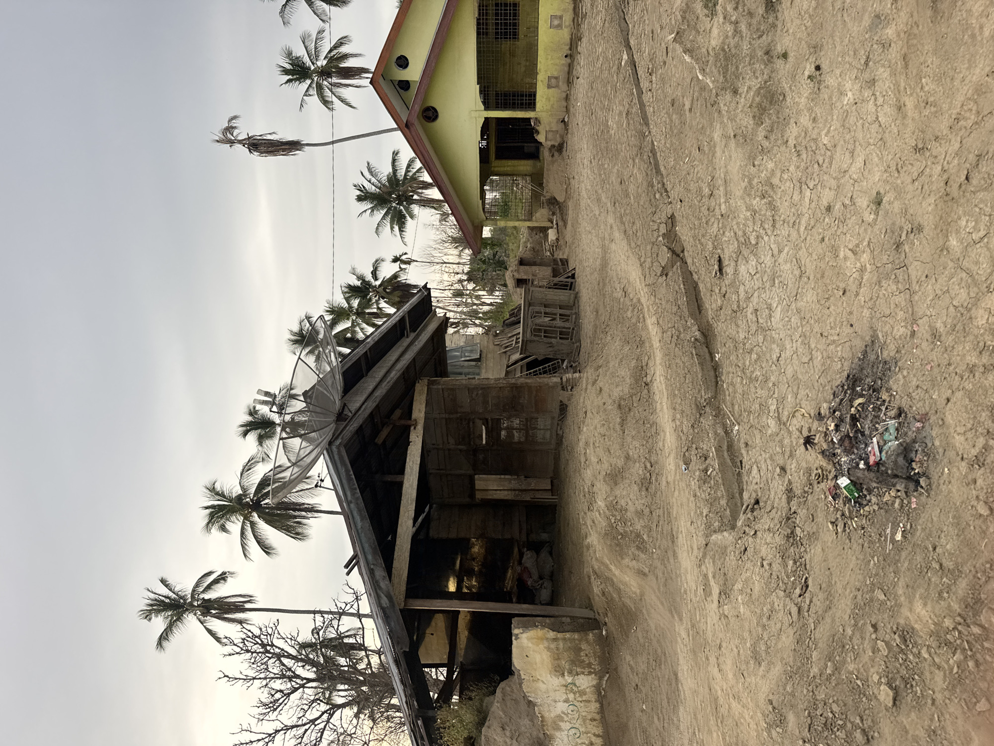 The image depicts a scene in Meunasah Raya, Indonesia, characterized by a dry, cracked earth ground. Two structures are visible. On the left is a dilapidated wooden shack with a corrugated metal roof, open on one side, revealing stored items. A satellite dish is mounted on its roof. To the right, a more intact, yellow painted house with a red-trimmed roof stands. Palm trees are scattered throughout the landscape. The sky is overcast. In the foreground, a pile of trash is visible on the cracked earth.