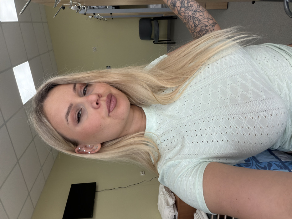 This image captures a blonde female taking a selfie in an indoor communal living space, likely a barracks or dormitory, in Sarapul, Russia. The female is positioned centrally, gazing directly into the camera with a neutral expression. She possesses long, straight, light blonde hair. Her left ear is adorned with multiple piercings, including several small studs and a star-shaped stud. She wears a light mint green, short-sleeved top featuring a mock neck and a textured pattern of small perforations. Her right forearm and upper arm are covered by a prominent black-ink tattoo depicting floral or mandala-like patterns. The room features light green walls and a white dropped ceiling with square fluorescent lighting panels. In the background to the left, a dark flat-screen television is mounted on the wall, with a white electrical outlet beneath it. To the right, metal frames, indicative of bunk beds or storage units, are visible, alongside a single dark grey utilitarian chair. Folded white linens are partially visible on a surface to the far left.