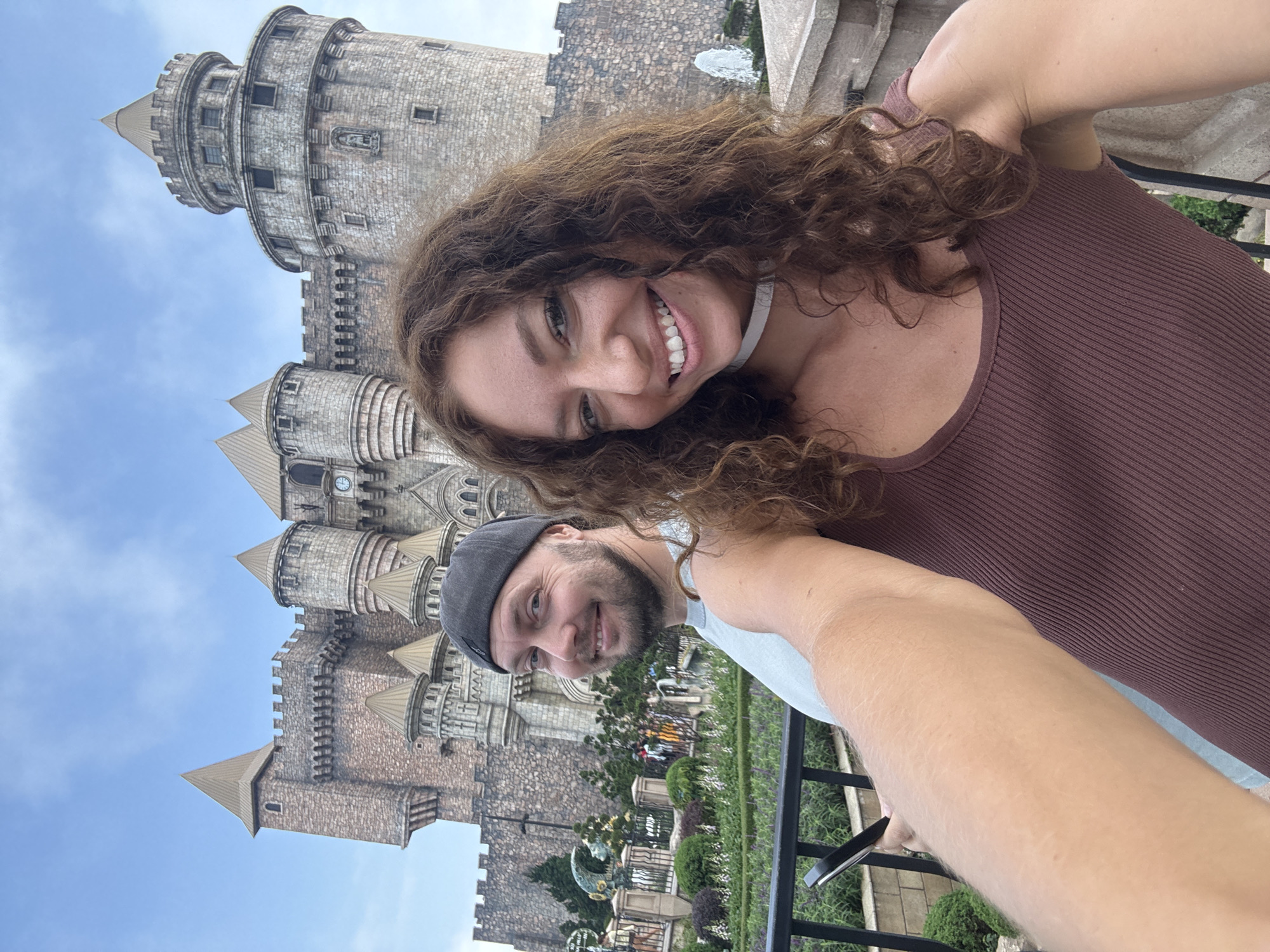 A selfie captures a smiling woman and man positioned in front of a majestic, multi-turreted stone castle. The woman, with long, curly brown hair and a wide smile, is closer to the camera, wearing a dark reddish-brown ribbed tank top. Beside her, a man with a short beard and a friendly smile wears a light blue t-shirt and a dark baseball cap turned backward.
The backdrop is dominated by the elaborate castle structure, built from various shades of grey and brown stone. It features numerous conical-roofed turrets, battlements, and windows, with a clock visible on one of the towers. The sky above is a clear blue with sparse white clouds, indicating daytime and pleasant weather. A black metal railing runs across the midground, separating the subjects from the castle, with glimpses of green foliage and sculpted bushes visible behind it.
Both individuals appear joyful and relaxed, suggesting they are enjoying their outing. The grand, European-inspired castle architecture strongly suggests the location is a notable tourist destination, consistent with places like Ba Na Hills in Đà Nẵng, Vietnam, where such structures are prominent. No visible text is discernible in the image.