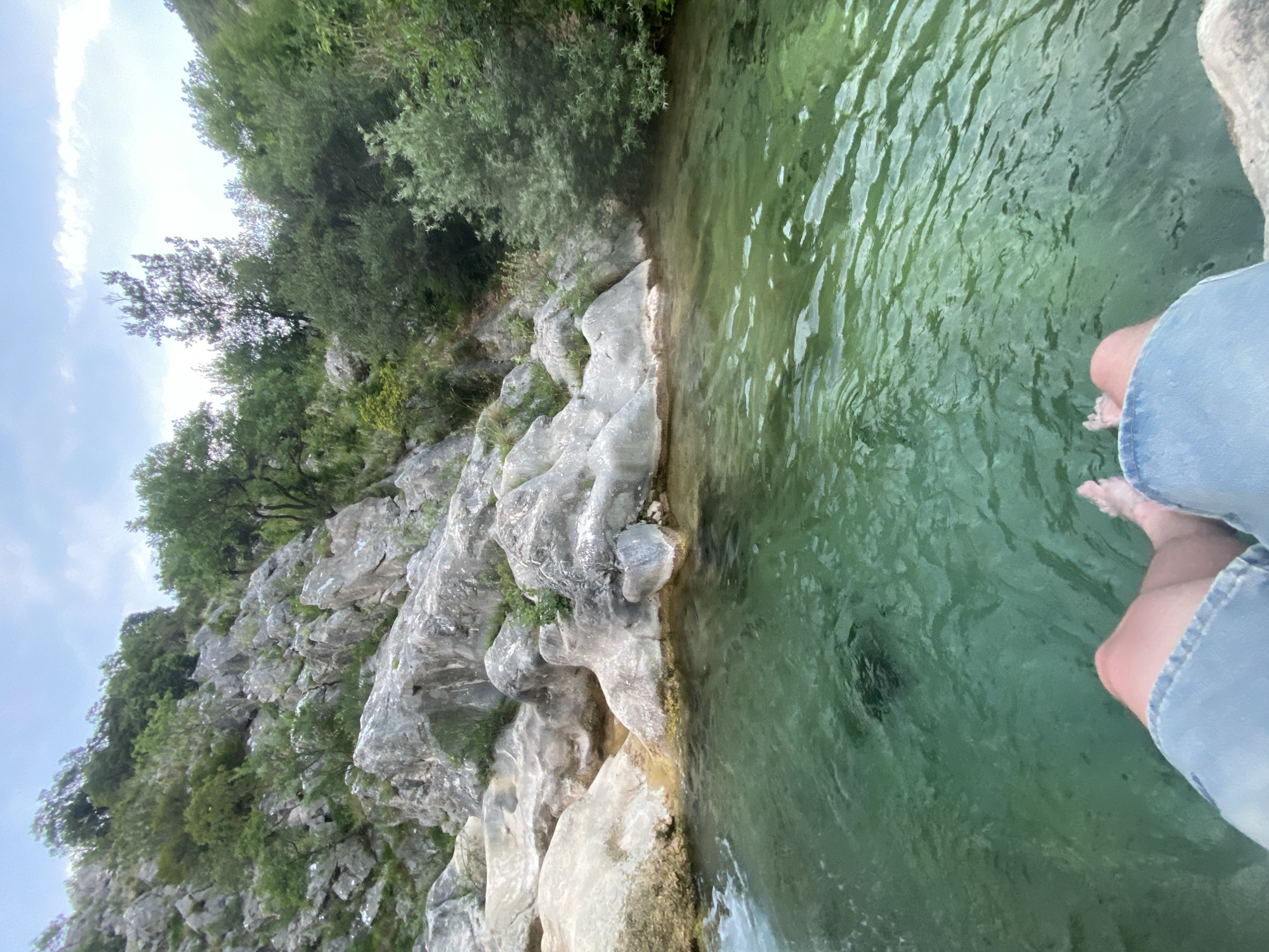 The image captures a serene natural scene, likely in Zupci, Montenegro, showing a person relaxing at the edge of clear, emerald-green water. From a slightly elevated perspective, the camera looks down towards the water and across to a rocky bank.
In the bottom right corner, the lower legs and bare feet of a person are visible, partially submerged in the cool, rippling water. The person appears to be wearing light blue denim shorts.
The right side of the frame is dominated by the tranquil water, which has a distinct green tint, suggesting purity and depth. Small ripples create patterns on its surface. The left side of the image features a rugged, rocky bank composed of light grey and white rock formations, likely limestone, with irregular, weathered textures. Sparse green vegetation, including small bushes and grass, grows in crevices and on ledges of the rocks.
Beyond the immediate rocky bank, a dense canopy of green trees stretches across the upper portion of the image, contributing to the lush natural environment. Above the trees, a bright sky with patches of white clouds is visible, indicating daytime, possibly mid-morning or early afternoon, with pleasant weather.
The overall scene conveys a sense of peaceful relaxation and connection with nature, as the individual enjoys dipping their feet into the refreshing water amidst the scenic landscape. No text is visible in the image.
