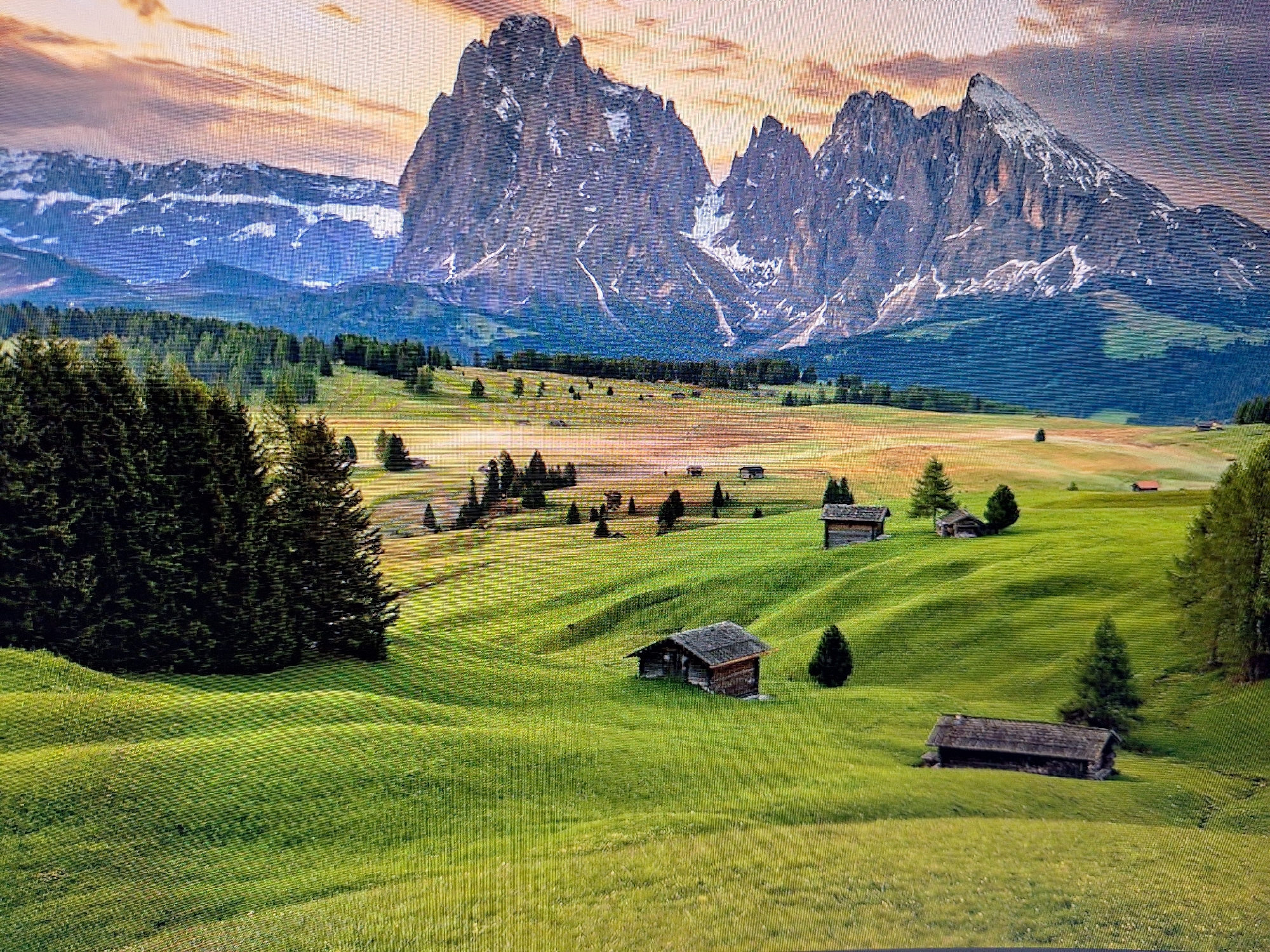 The image depicts a breathtaking mountain landscape with rolling green hills dotted with small wooden chalets. In the foreground, lush green fields stretch out, with gentle undulations creating a sense of depth. Several rustic wooden structures, likely mountain huts or barns, are scattered across the landscape, their dark roofs contrasting with the vibrant greenery.
In the middle ground, a band of dark green coniferous trees forms a dense forest line, separating the lower hills from the dramatic mountain range that dominates the background. These mountains are jagged and imposing, with snow-capped peaks reaching towards a sky filled with warm, sunset hues of orange, pink, and purple. The interplay of light and shadow on the mountain faces highlights their rugged texture.
The scene is one of serene natural beauty, evoking a feeling of tranquility and remoteness. There are no people or animals visible in the image. The environment is characterized by its natural elements: mountains, trees, and open fields, suggesting a rural or alpine setting. The time of day appears to be late afternoon or early evening, given the warm, diffused light and the colors in the sky.
This image is not of Varna, Bulgaria. Varna is a coastal city known for its beaches and Black Sea views, which are not represented in this mountainous, inland landscape.