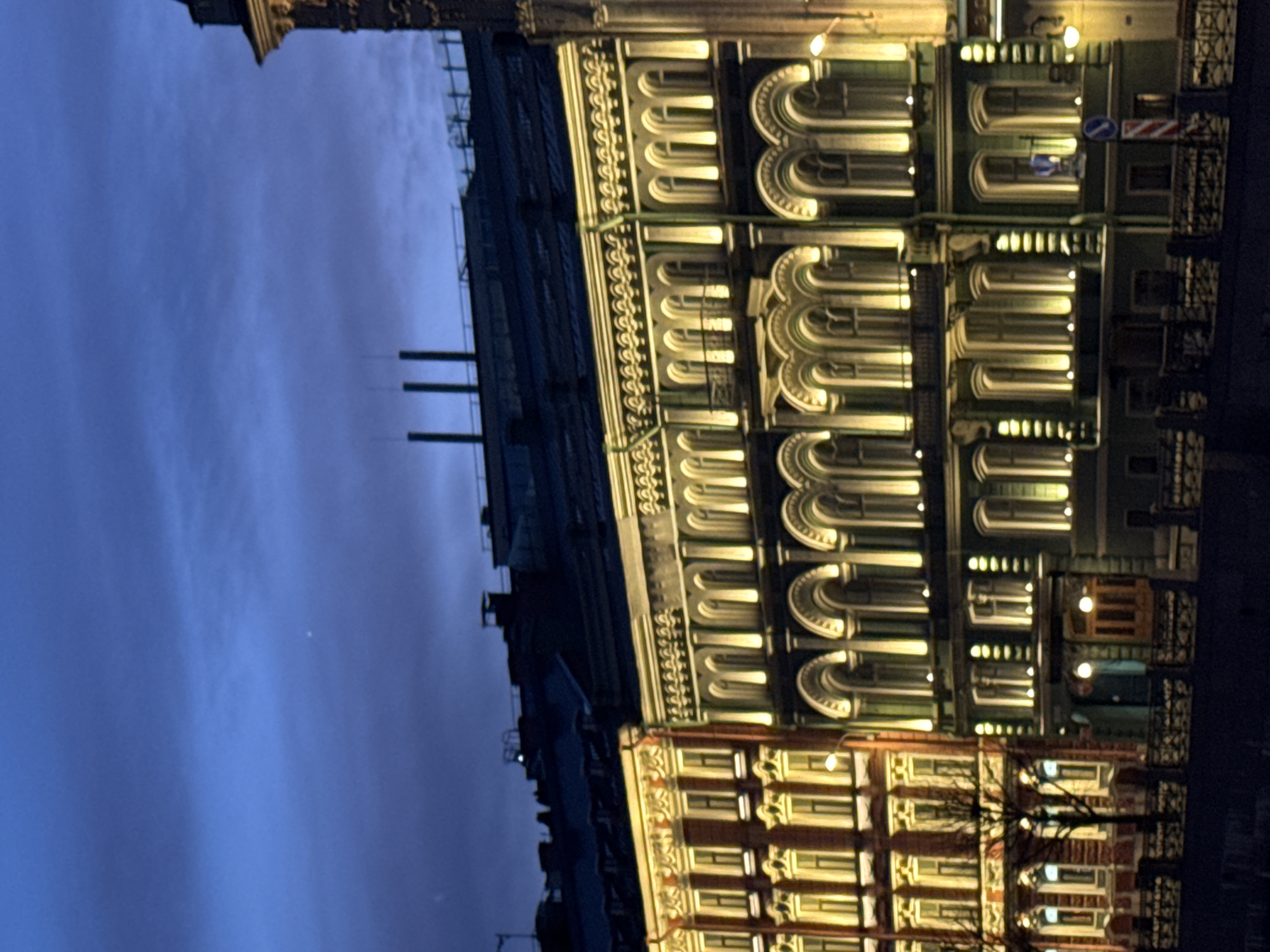 A nighttime image shows a building in St. Petersburg, Russia, with illuminated windows and ornate architectural details. The sky is a dark blue with some clouds. The building is multi-storied, with a mix of architectural styles in the different levels. Street lights are visible.