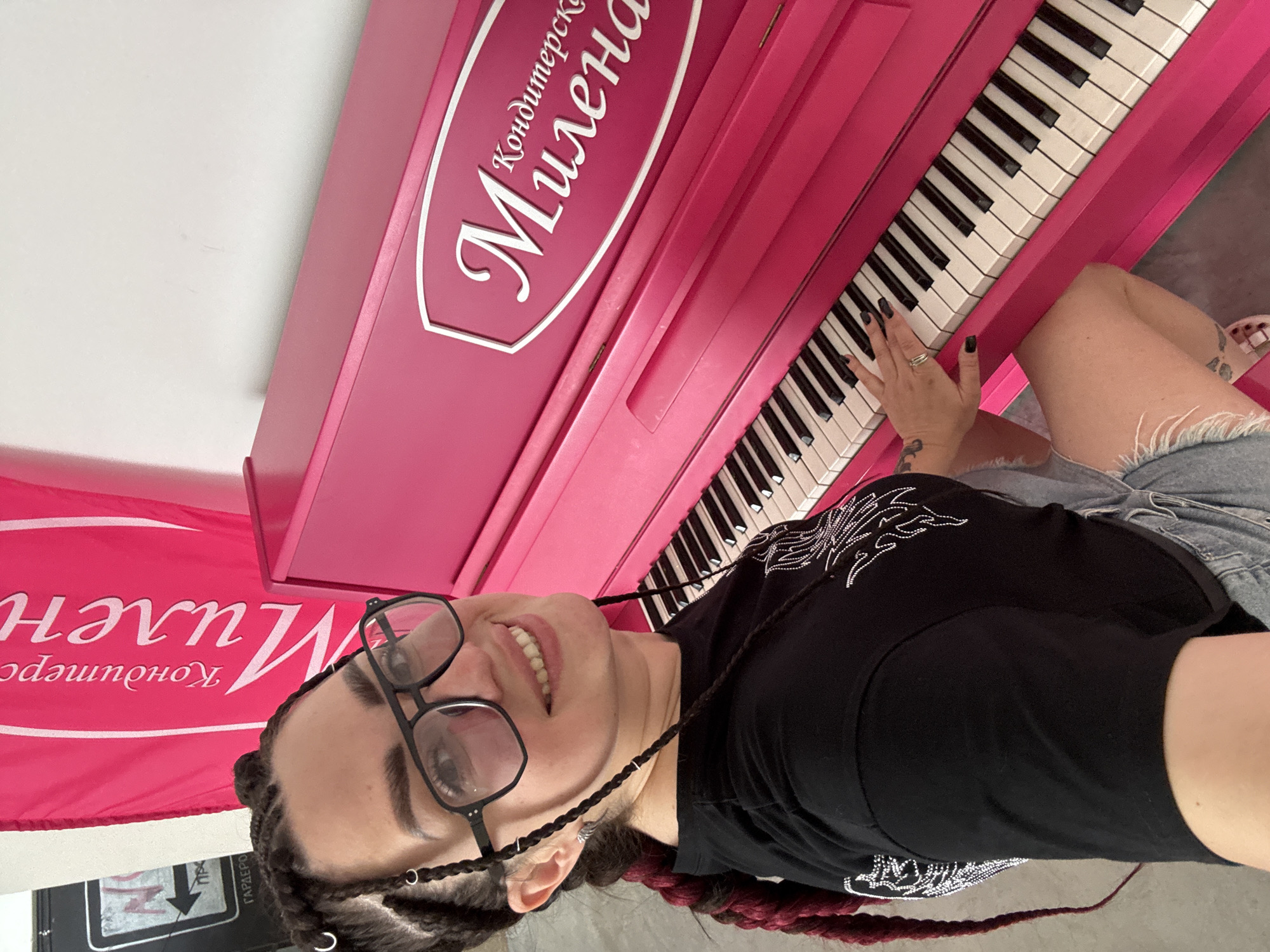 A woman with dark braided hair, wearing glasses, a black t-shirt, and denim shorts, sits in front of a bright pink piano, with her right hand on the keys. A pink banner with white text, "Кондитерска Милена," hangs behind her to the left. To the left of the banner, a black sign with white text and an arrow is visible. The background is a plain white wall. The woman is smiling and looking towards the camera.