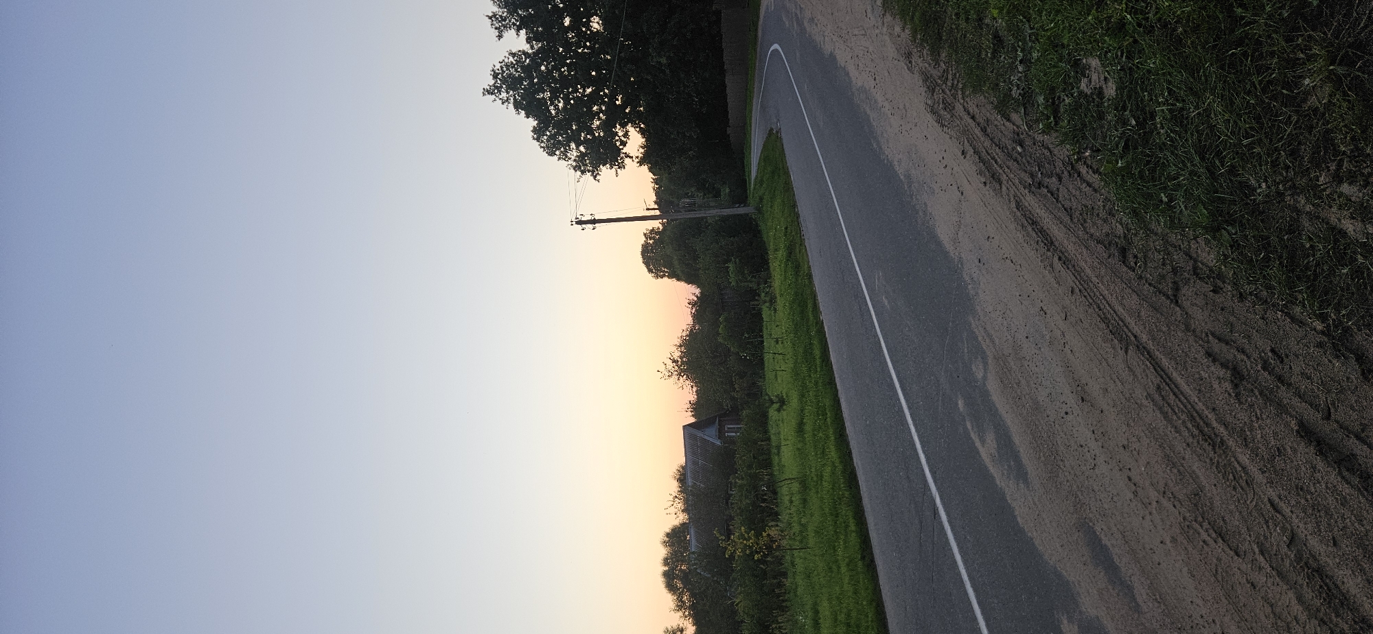 The image depicts a rural scene in Мазалаўскі сельскі Савет, Belarus, during what appears to be either sunrise or sunset. A paved road with a white line marking curves to the right. The road is bordered by a dirt shoulder with visible tire tracks and patches of grass. On the left side of the road, a green grassy area with some small trees leads up to a cluster of trees and a building with a grey, sloped roof. On the right side of the road, a wooden utility pole with wires extends upwards, and behind it, a large, dark green tree dominates the background, with more trees and a fence visible in the distance. The sky above is a gradient of pale blue to a soft yellow-orange hue near the horizon, suggesting twilight. The overall atmosphere is serene and quiet.