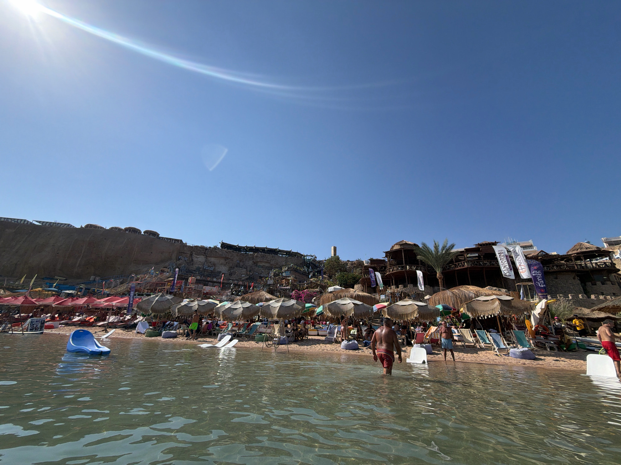 The image depicts a vibrant beach scene in Sharm El Sheikh, Egypt, likely during daylight hours given the bright sun and clear blue sky. The foreground shows clear, shallow water with ripples reflecting the sunlight. A blue inflatable slide is positioned in the water, inviting use.
The beach itself is populated with numerous beach umbrellas, providing shade for sunbathers. Beach chairs are visible beneath the umbrellas, and people are scattered across the sand, some sitting, some standing, and some walking. The beach appears to be backed by a rocky incline with buildings and structures built into the hillside, giving a sense of a resort or entertainment complex. Flags with various logos are displayed on some of the buildings.
Several individuals are visible in the water and on the beach, all appearing to be casually dressed for a beach environment, mostly in swimsuits. One man is walking out of the water towards the beach. The overall atmosphere is one of leisure and recreation. The architecture and the lush vegetation (including palm trees) suggest a warm, possibly tropical or subtropical climate, consistent with Sharm El Sheikh. The presence of sunbathers, umbrellas, and beach equipment confirms it is a popular tourist destination.