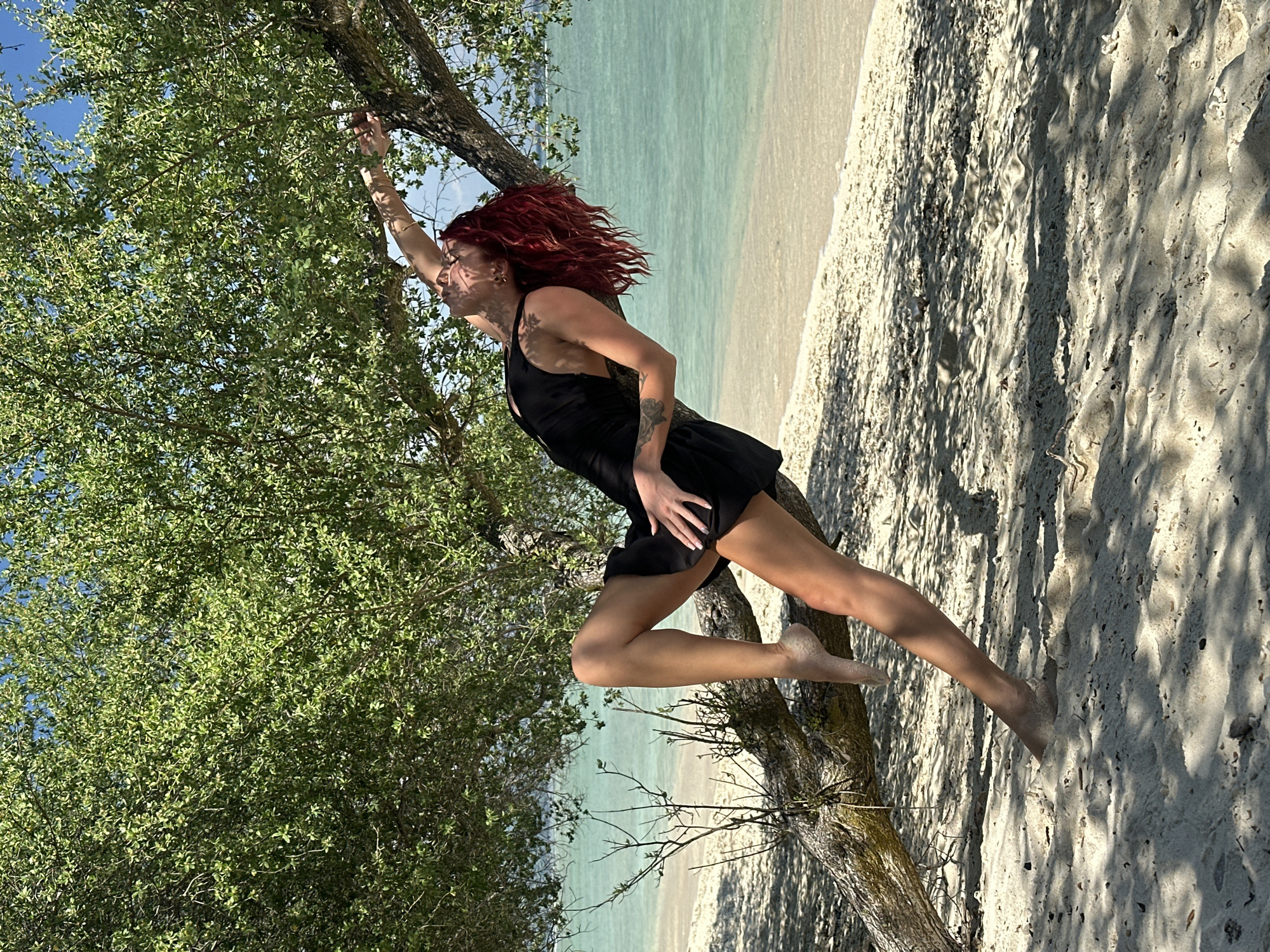 A woman with red hair is standing on a tree branch on a beach, with a black dress on. The tree is surrounded by green foliage, and the beach has white sand and blue-green water. The location appears to be in Unknown, Indonesia.