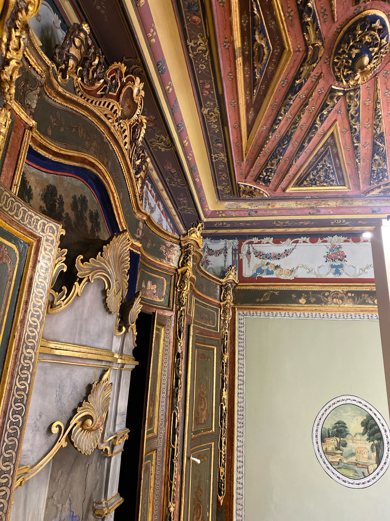 The image shows the interior of a richly decorated room, likely a historic building. The ceiling is ornate, featuring geometric patterns and intricate gold detailing with pink and blue accents. The walls are adorned with elaborate painted panels, including floral motifs and landscape scenes within circular frames. The architecture itself is highly decorative, with gilded carved elements and colorful painted surfaces framing doorways and architectural features. The room appears to be part of a palace or a grand residence, possibly in the city of Sarıyer, Turkey, given the style of decoration often found in historical Ottoman architecture. There are no people visible in the image, and the scene depicts an static, empty interior.