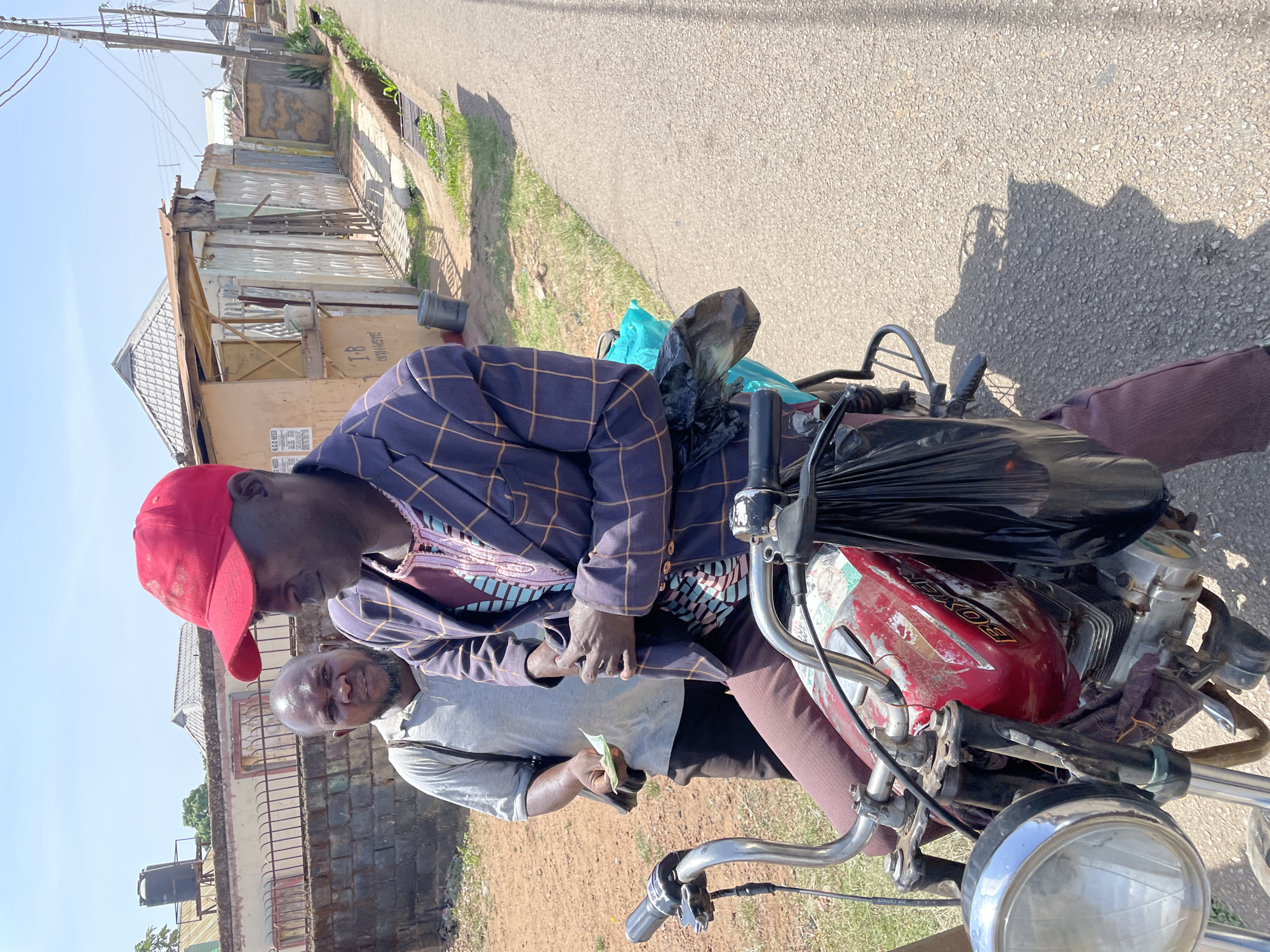 In Jos, Nigeria, a man wearing a red cap and a blue plaid blazer is seated on a red motorcycle, possibly conducting a transaction with another man in a grey shirt holding a green bill. The location appears to be a residential street with buildings and vegetation visible in the background.