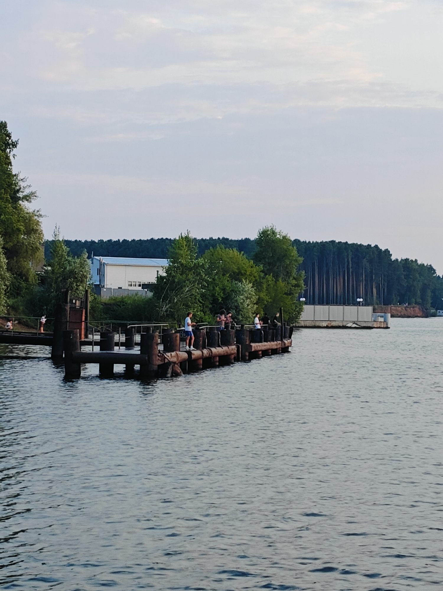 The image depicts a wide body of water, likely a river or lake, with a wooden pier extending into the foreground. Several individuals are visible on the pier, some standing and observing, while others appear to be engaged in casual conversation or activity. The pier itself is weathered and appears to be made of thick wooden pilings and planks.
In the background, the scene transitions to a densely wooded area with tall, slender trees forming a dark green line against the horizon. Behind the trees, a light-colored building with a blue roof is partially visible, suggesting human habitation or infrastructure near the natural landscape. The sky above is partly cloudy, with soft, diffused light indicating it might be late afternoon or early evening. The overall atmosphere is serene and relaxed, with the gentle ripples on the water adding to the tranquility. The presence of people on the pier suggests it's a place for leisure or perhaps a point of transit. The location is identified as Chaikovsky, Russia. There is no visible text in the image.
