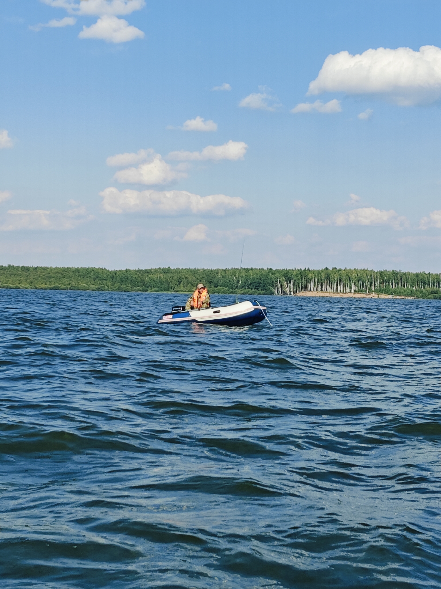 A man wearing an orange life vest sits in a blue and white inflatable boat on a dark blue, rippling lake. He is facing away from the camera, looking towards a distant, tree-lined shore. A fishing rod is visible, sticking up from the boat. The sky is a bright blue with scattered white clouds. The scene appears to be a natural, outdoor setting, possibly a reservoir or large lake, in Chaykovsky, Russia.