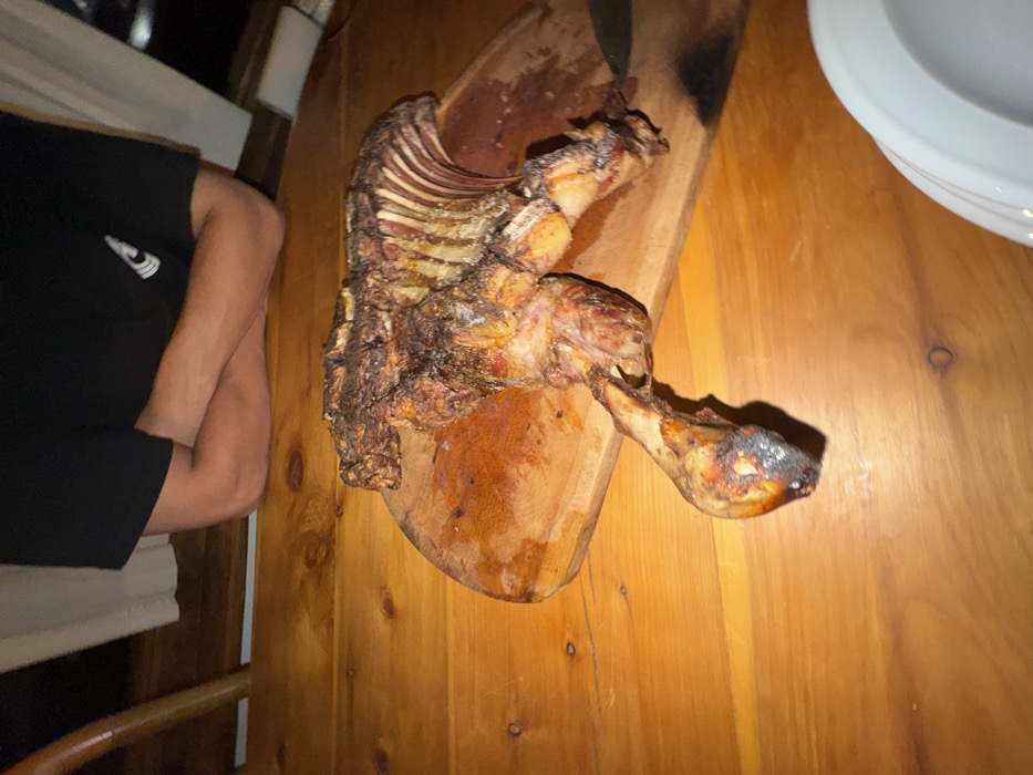 A cooked lamb carcass sits on a wooden cutting board. The lamb appears heavily seasoned and roasted, with the rib cage and leg bones exposed. The board rests on a wooden table, accompanied by a plate and a person in a black t-shirt. The scene is indoors, potentially a home dining setting.