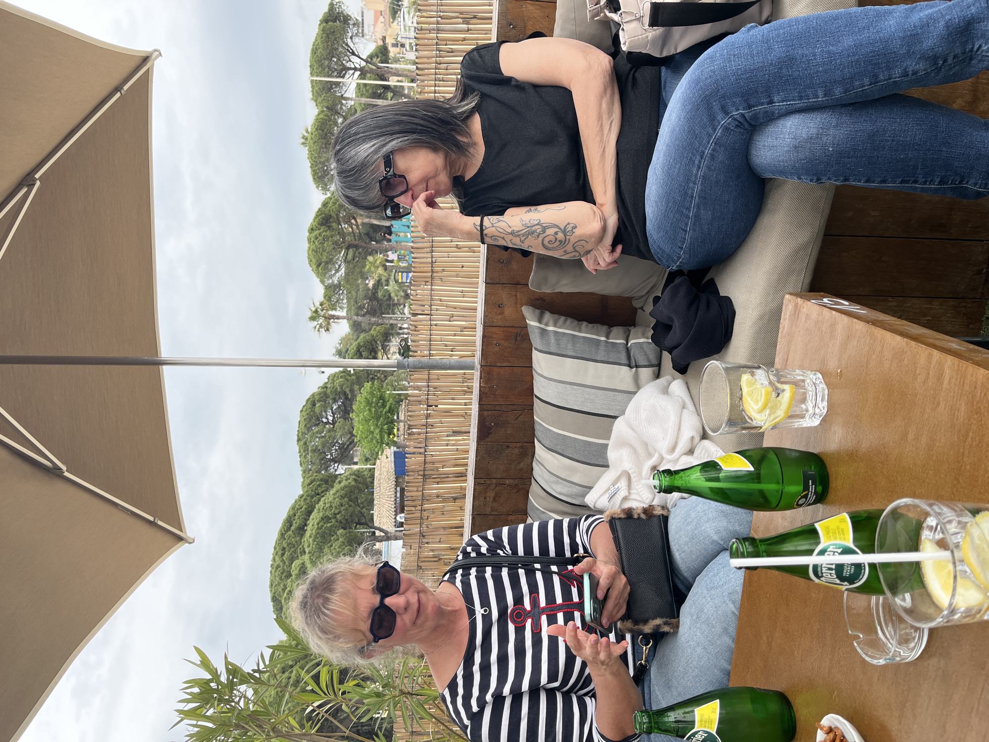 Two middle-aged women are seated on a cushioned bench next to a wooden table at an outdoor cafe in Sainte-Maxime, France. One woman, with short blonde hair and sunglasses, wears a black and white striped shirt with a red anchor design and blue jeans. She is holding a smartphone and a black purse. The second woman, with gray hair, sunglasses, and a tattoo on her left forearm, is wearing a black t-shirt and blue jeans. The table holds two green glass bottles of Perrier, two glasses with lemon wedges, a bowl of snacks, and a white straw. An umbrella is partially visible, providing shade. A fence and trees are in the background.