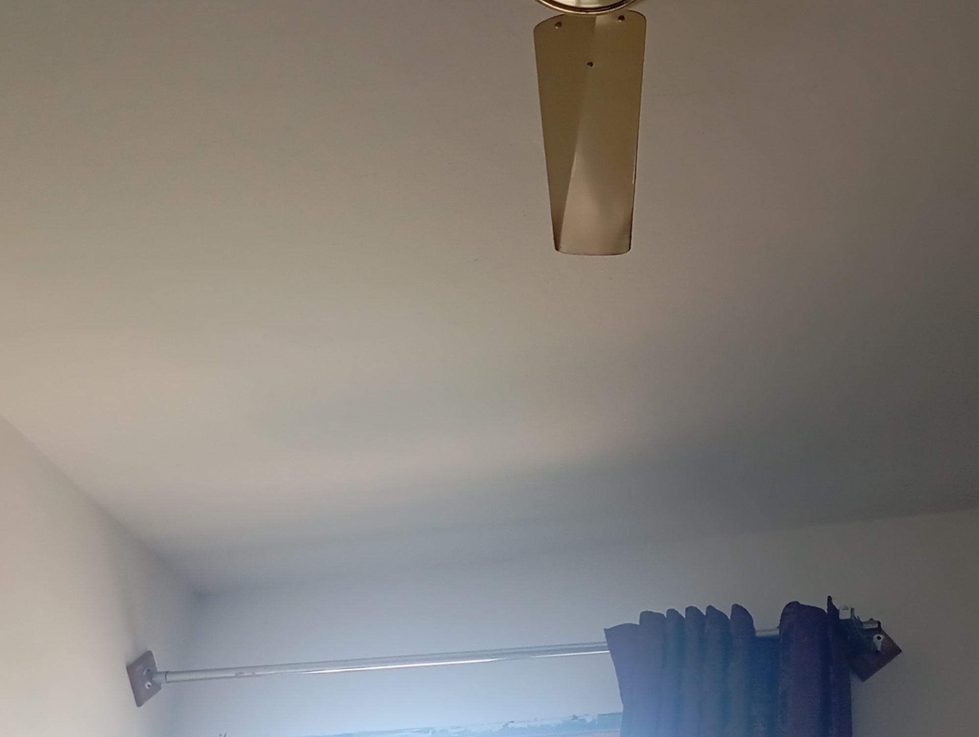 The image captures an indoor scene, likely a room, with a ceiling fan blade visible at the top center. The fan blade is a metallic, angled shape. Below, a curtain rod stretches horizontally across the frame, with a portion of dark purple curtains drawn to the right. A white wall is visible on the left side of the image, meeting the ceiling. There is no indication of the city of Lassan, Pakistan, or any specific location details within the image itself. The scene appears to be static, with no discernible actions or interactions taking place.