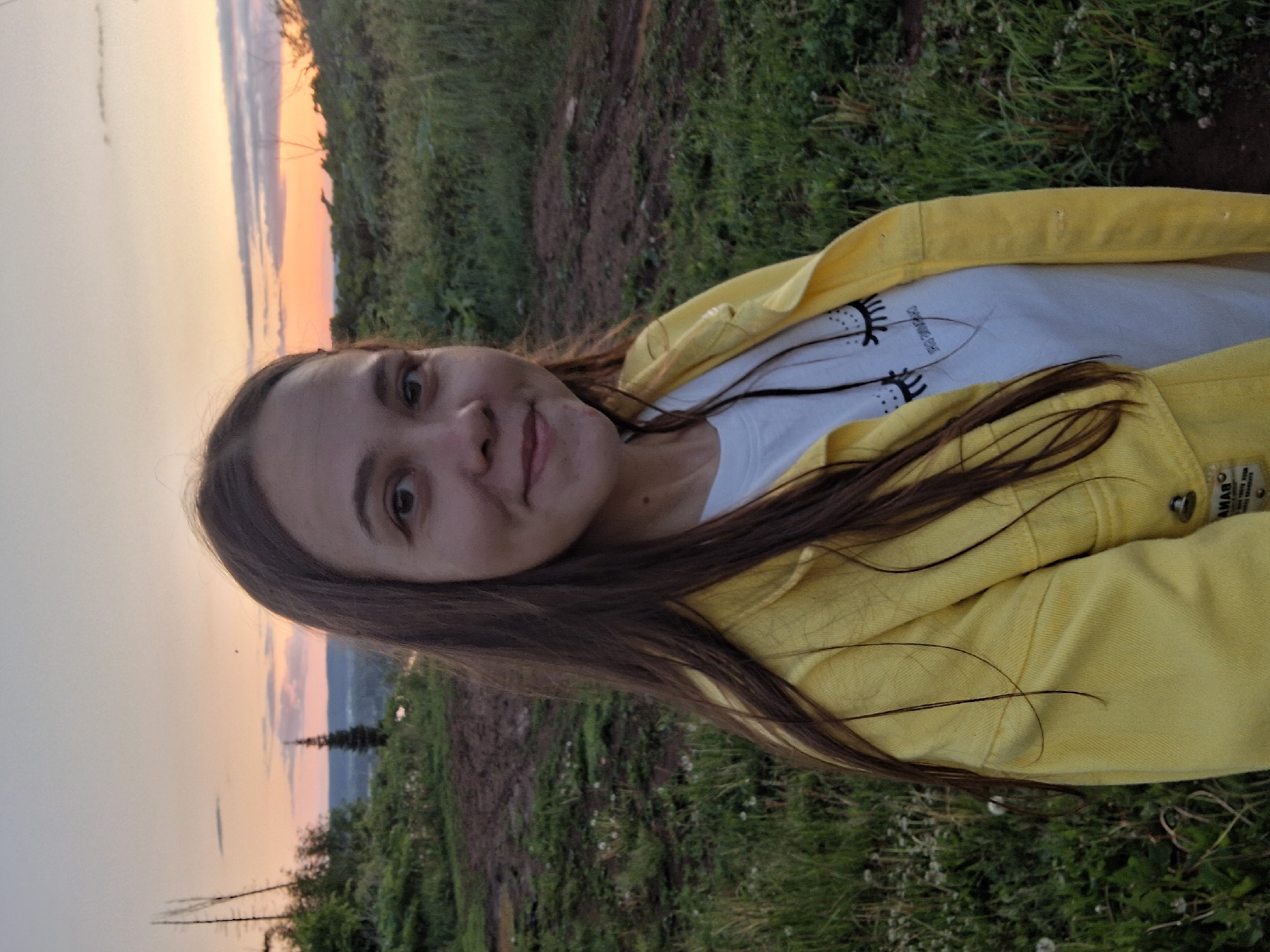 A young woman is captured in an outdoor setting at sunset, likely in the natural landscapes around Chaykovsky, Russia. She has long, dark brown hair and is looking directly at the camera with a soft, contented smile. She wears a bright yellow jacket over a white t-shirt that features a distinctive graphic design of stylized eyes with long black lashes.
The background showcases a picturesque sunset. The sky displays warm hues of orange and pink near the horizon, fading into lighter blue tones above. Distant hills or mountains and scattered trees are visible against the vibrant sky, suggesting the photo was taken from an elevated position overlooking the landscape. The foreground consists of green grass and patches of earth.
The scene conveys a calm and serene atmosphere, highlighting the beauty of nature during the golden hour. A label on the yellow jacket is partially visible, showing the text "AMABO" and "MADE IN".